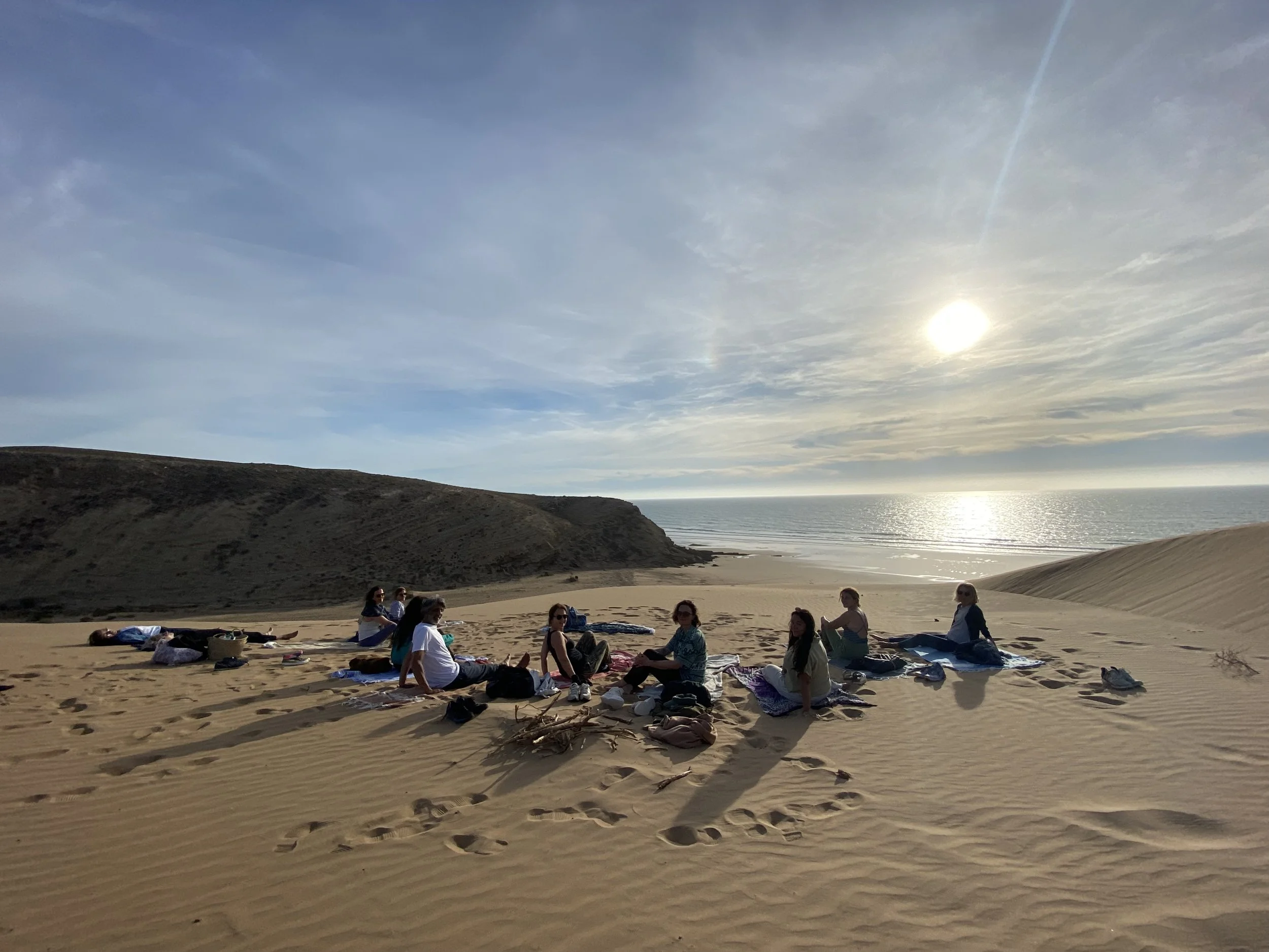 Groupe de personnes assises sur des dunes de sable face à la mer, sous un ciel ensoleillé.
