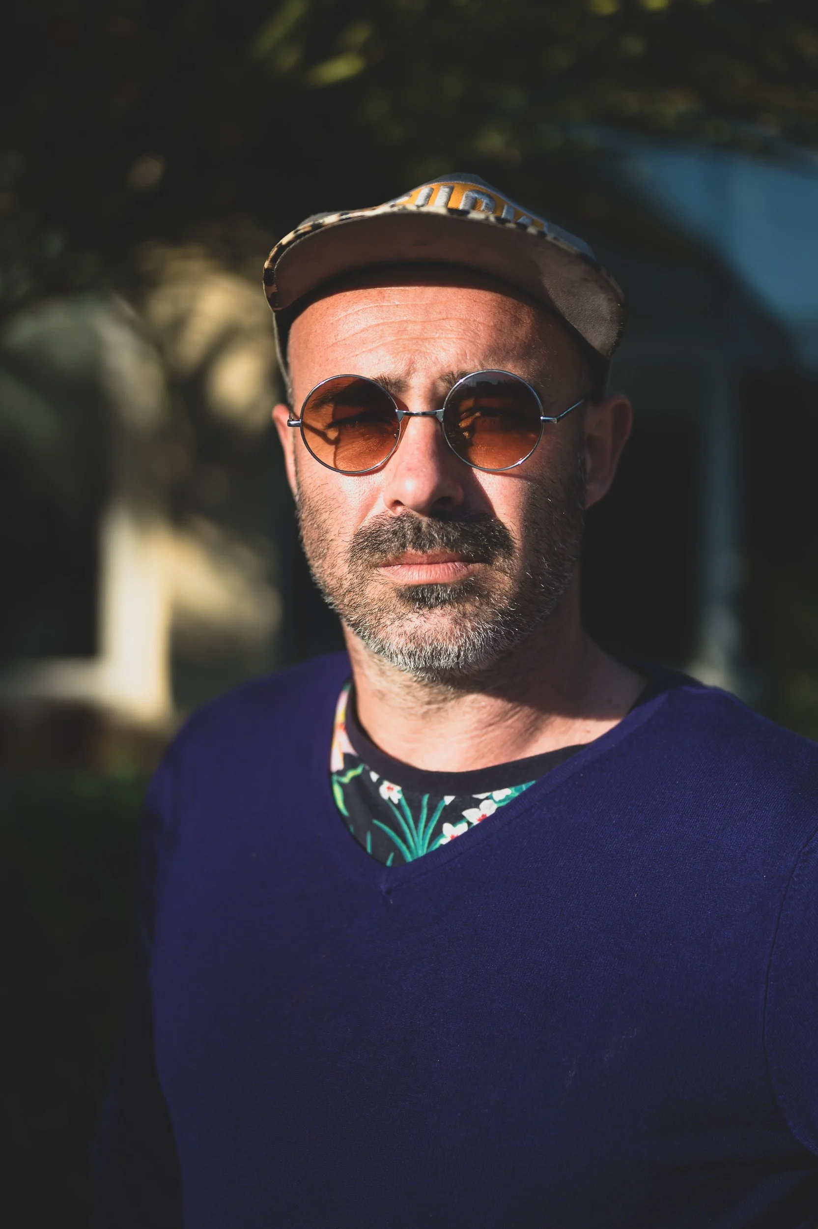 Un homme portant des lunettes de soleil rondes, un bonnet et un t-shirt bleu, posant en plein air avec un fond flou de nature.