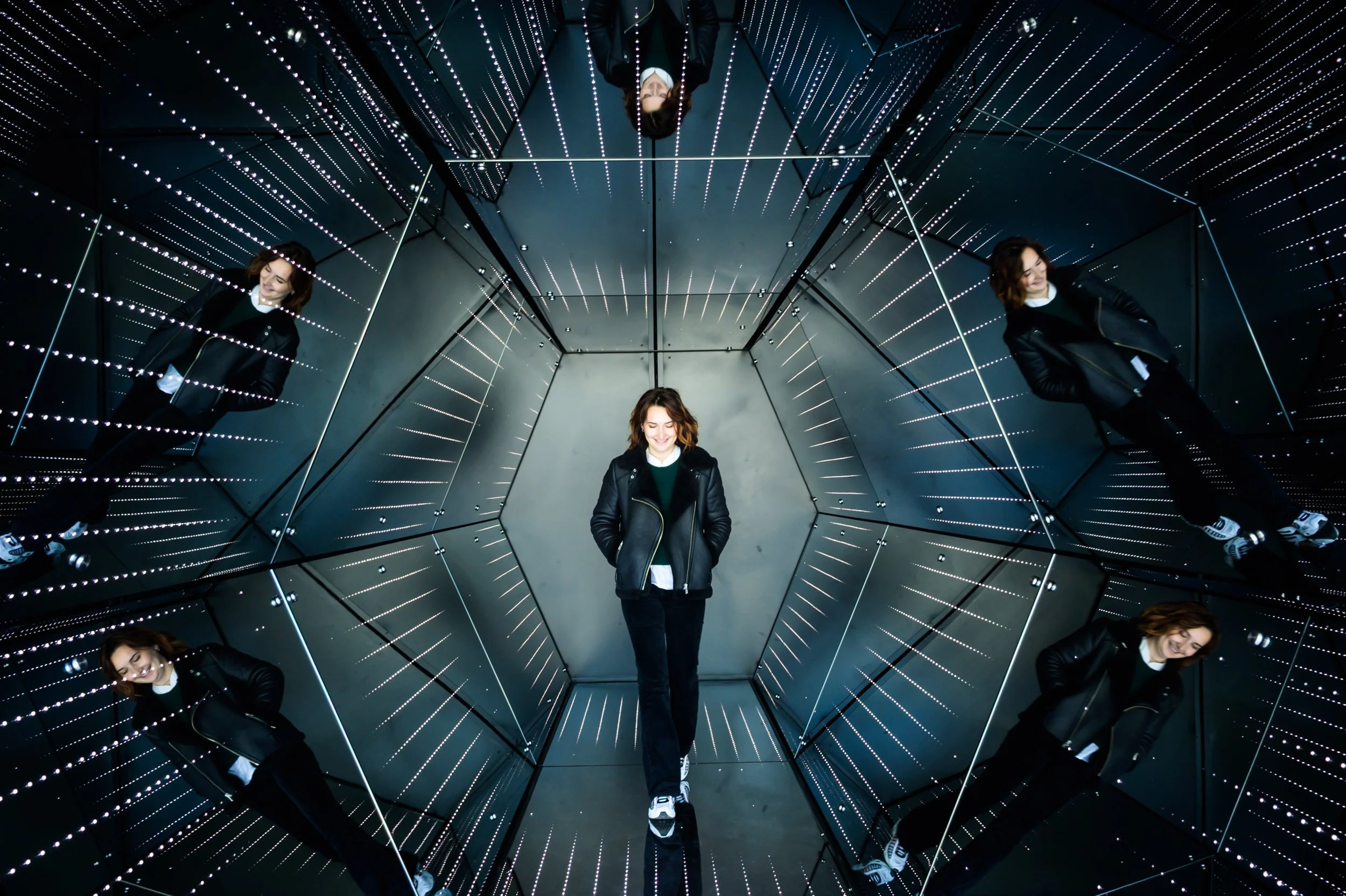 Une femme avec des cheveux marron court, portant un blouson en cuir noir, une chemise blanche et un pull vert, se tient debout dans un espace où des miroirs sont disposés de façon à créer une illusion de multiplication infinie d'elle-même grâce à des