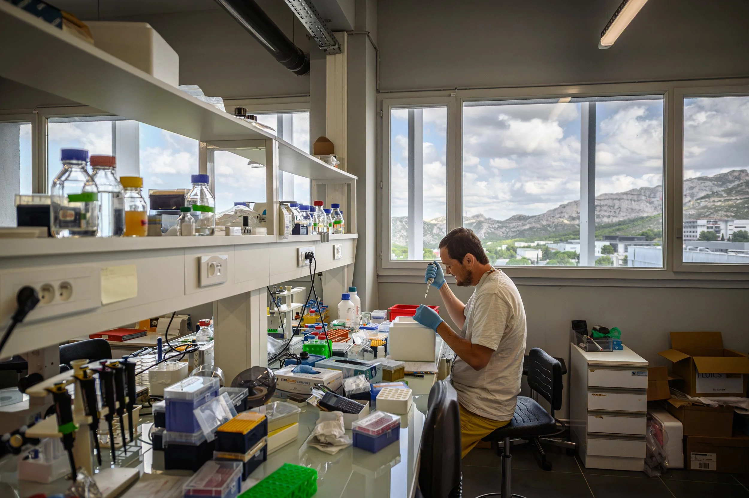 Un laboratoire avec un chercheur travaillant à un laboratoire, entouré de bocaux, de pipettes, d'ordinateurs et d'autres équipements scientifiques, avec de grandes fenêtres offrant une vue sur des montagnes et un ciel partiellement nuageux.
