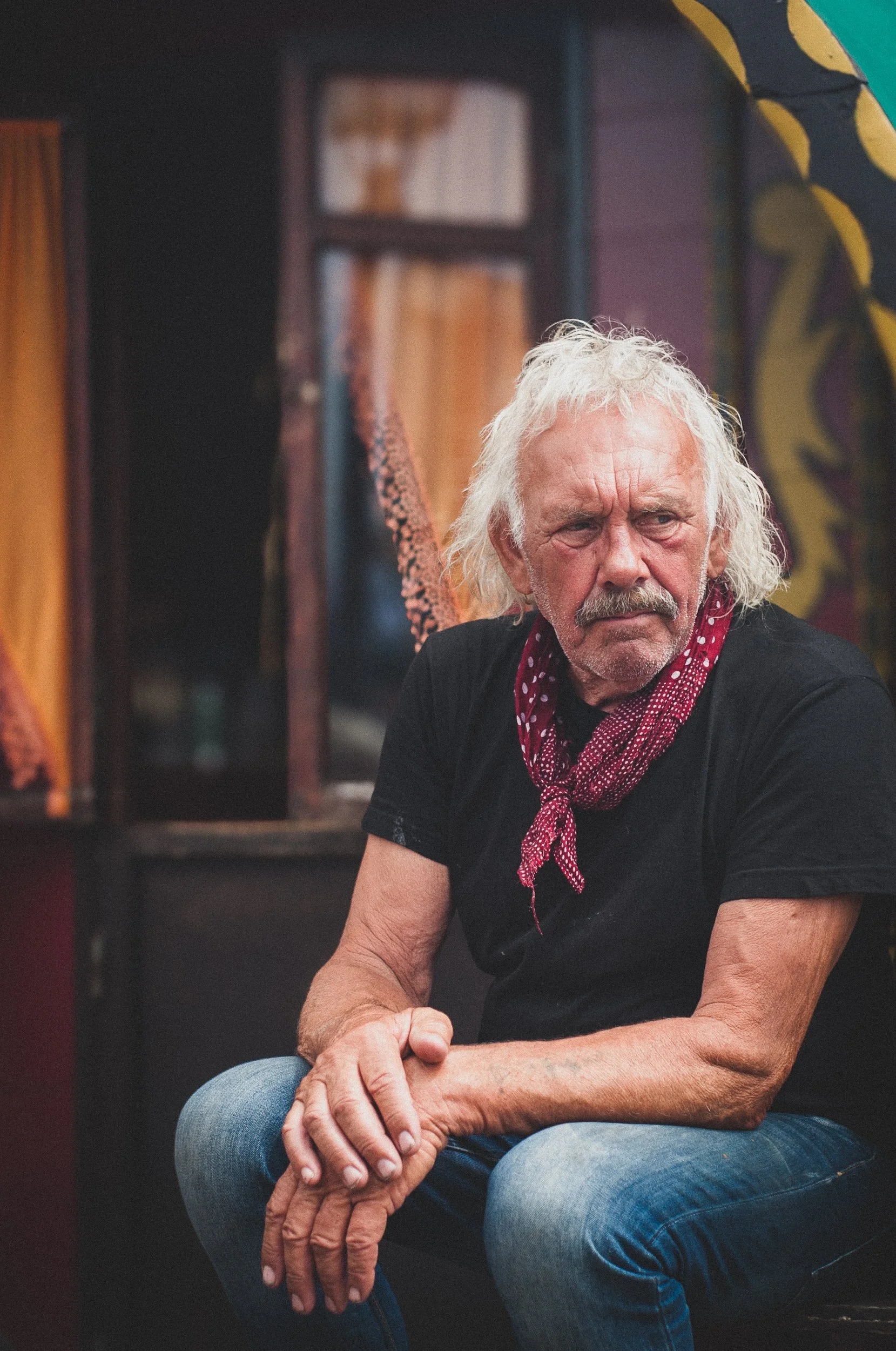 Un vieil homme aux cheveux blancs, portant un bandana rouge à pois, assis avec ses mains croisées, dans une pièce décorée de couleurs vives.