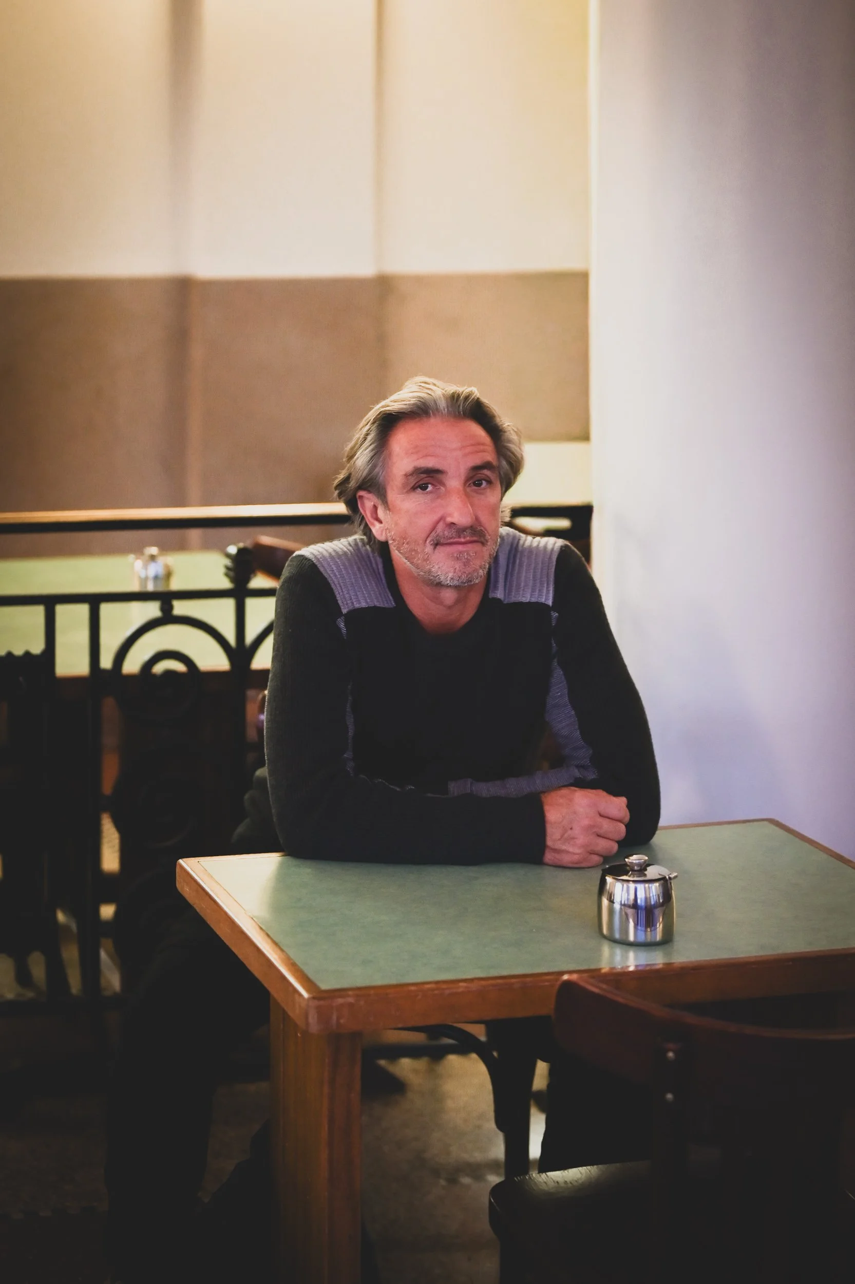 Un homme assis à une table dans un café, regardant vers l'appareil photo. La table a un petit pot en métal. L'arrière-plan est flou avec des murs beige et un garde-corps noir.