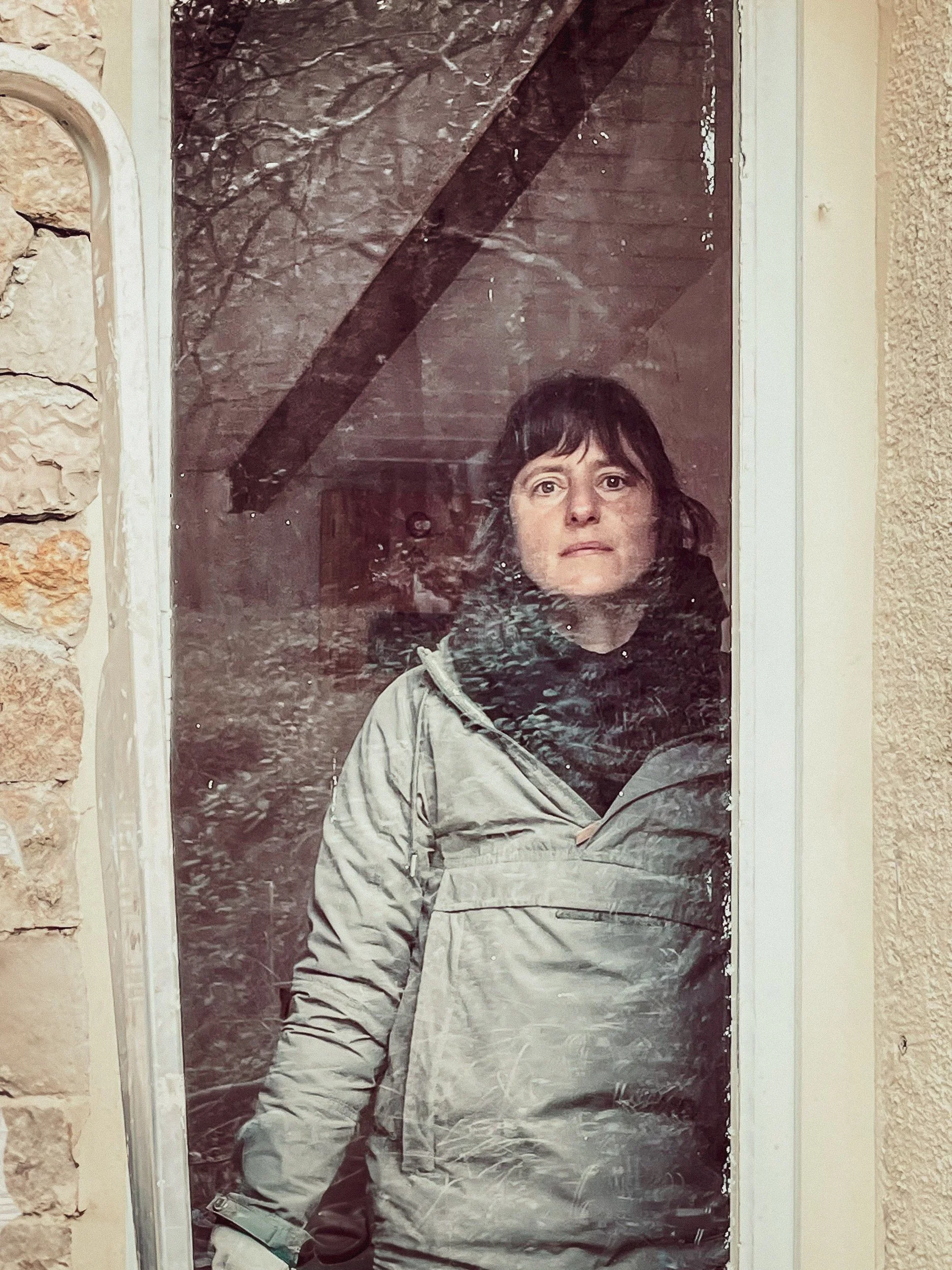 Une femme regarde à travers une vitre sale, portant une veste beige avec une capuche noire, dans un environnement intérieur ou extérieur brouillé par la saleté sur la vitre.