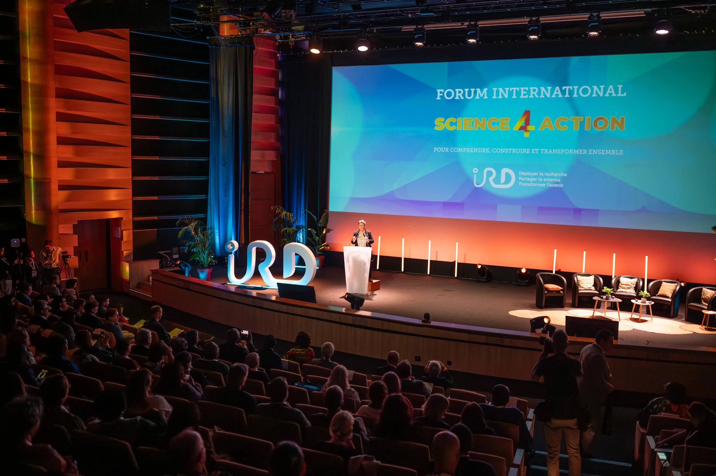 Une salle de conférence avec un écran géant affichant le texte 'Forum International Science 4 Action'. Des personnes assises regardent la présentation, et une femme est en train de parler à un pupitre sur scène.