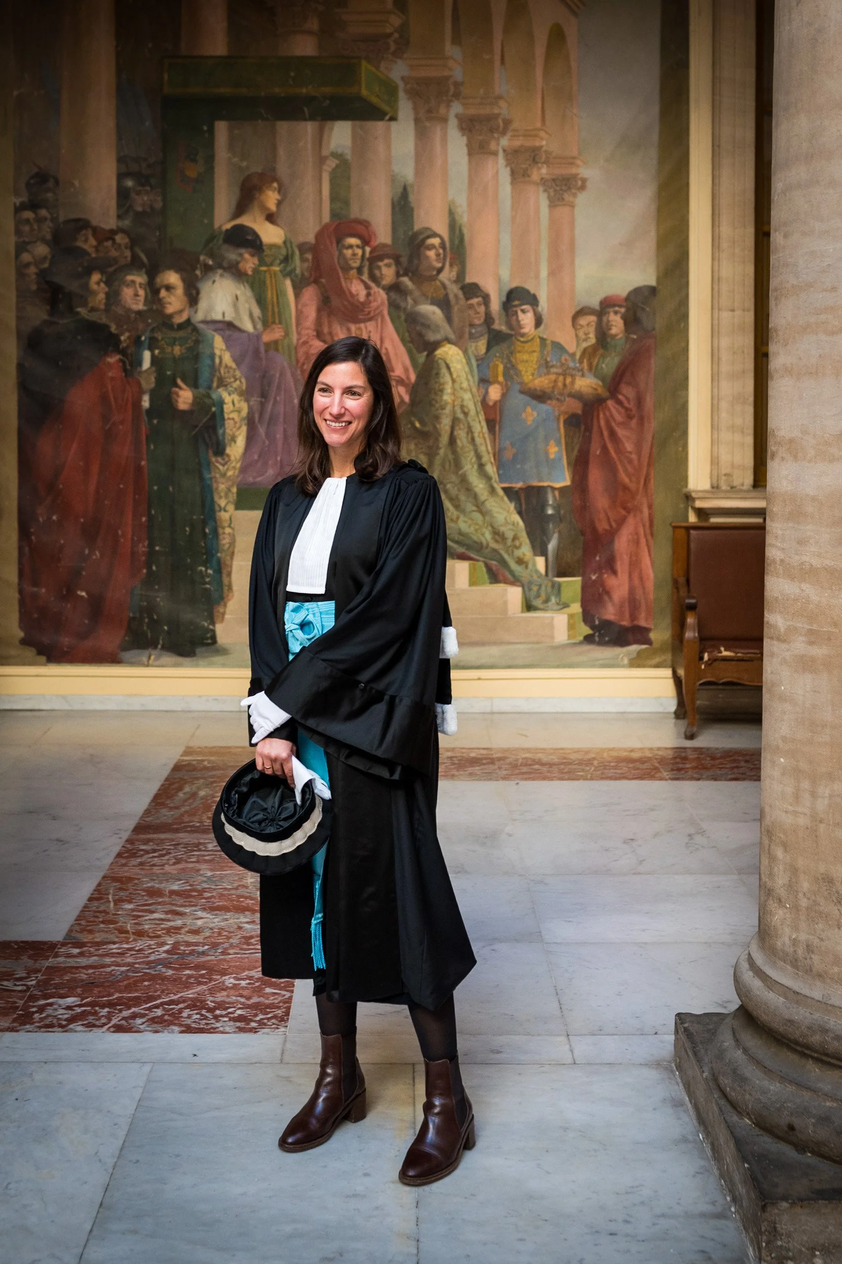 Une femme portant une robe de juge avec une toque, debout dans un musée, en face d'une grande peinture historique représentant une scène royale ou historique, entourée de colonnes de pierre et de murs en pierre.