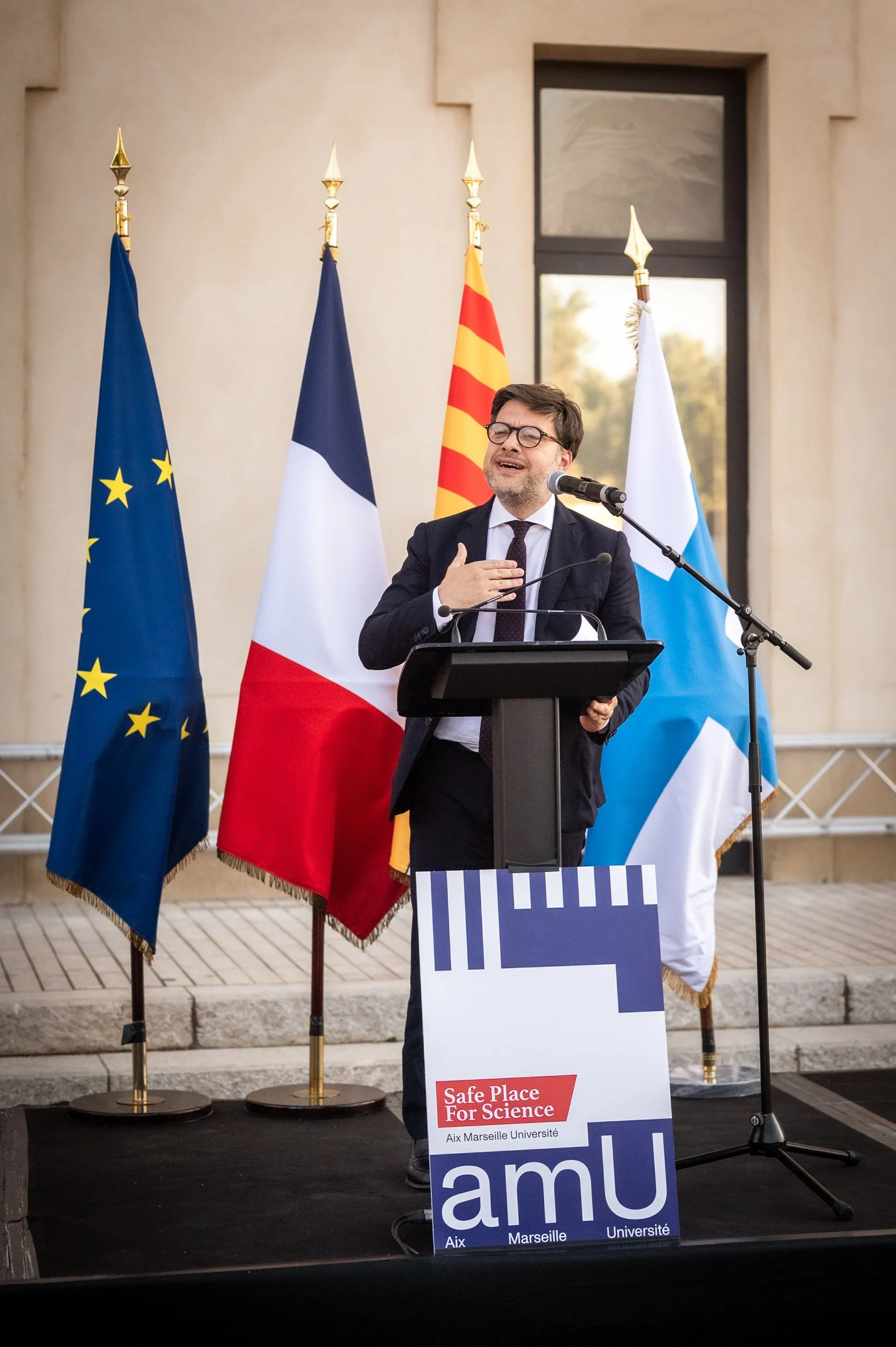 Un homme parle à un micro lors d'une conférence de presse en extérieur, derrière lui trois drapeaux (de l'Union européenne, de la France, de la Catalogne et de la Russie) sont visibles. Un panneau indique 'Safe Place for Science' et 'Aix Marseille Un