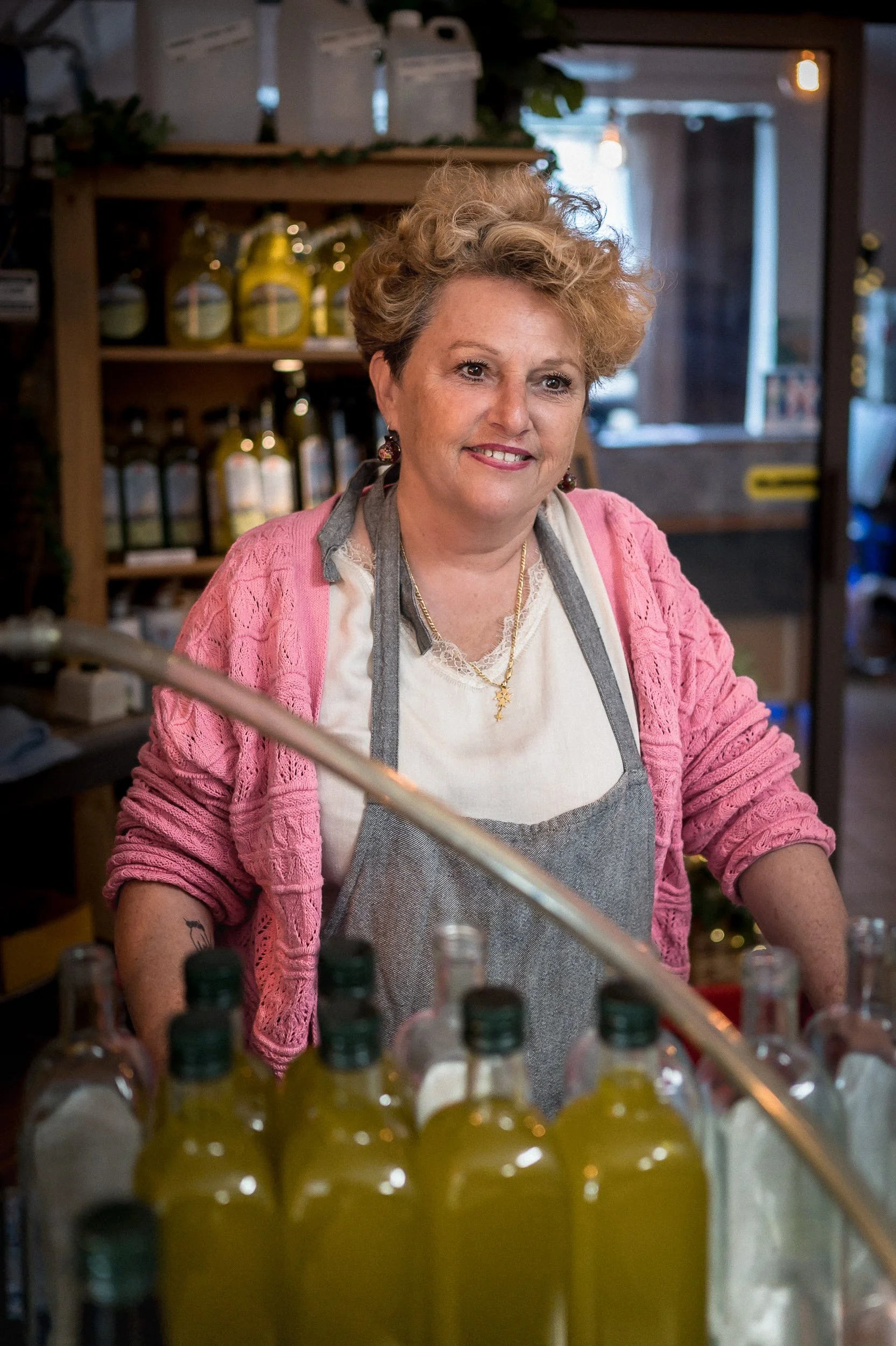 Une femme souriante portant un tablier gris et un cardigan rose, se trouve derrière un comptoir avec des bouteilles d'huile ou d'une autre substance, dans un intérieur chaleureux comme une épicerie ou un marché.