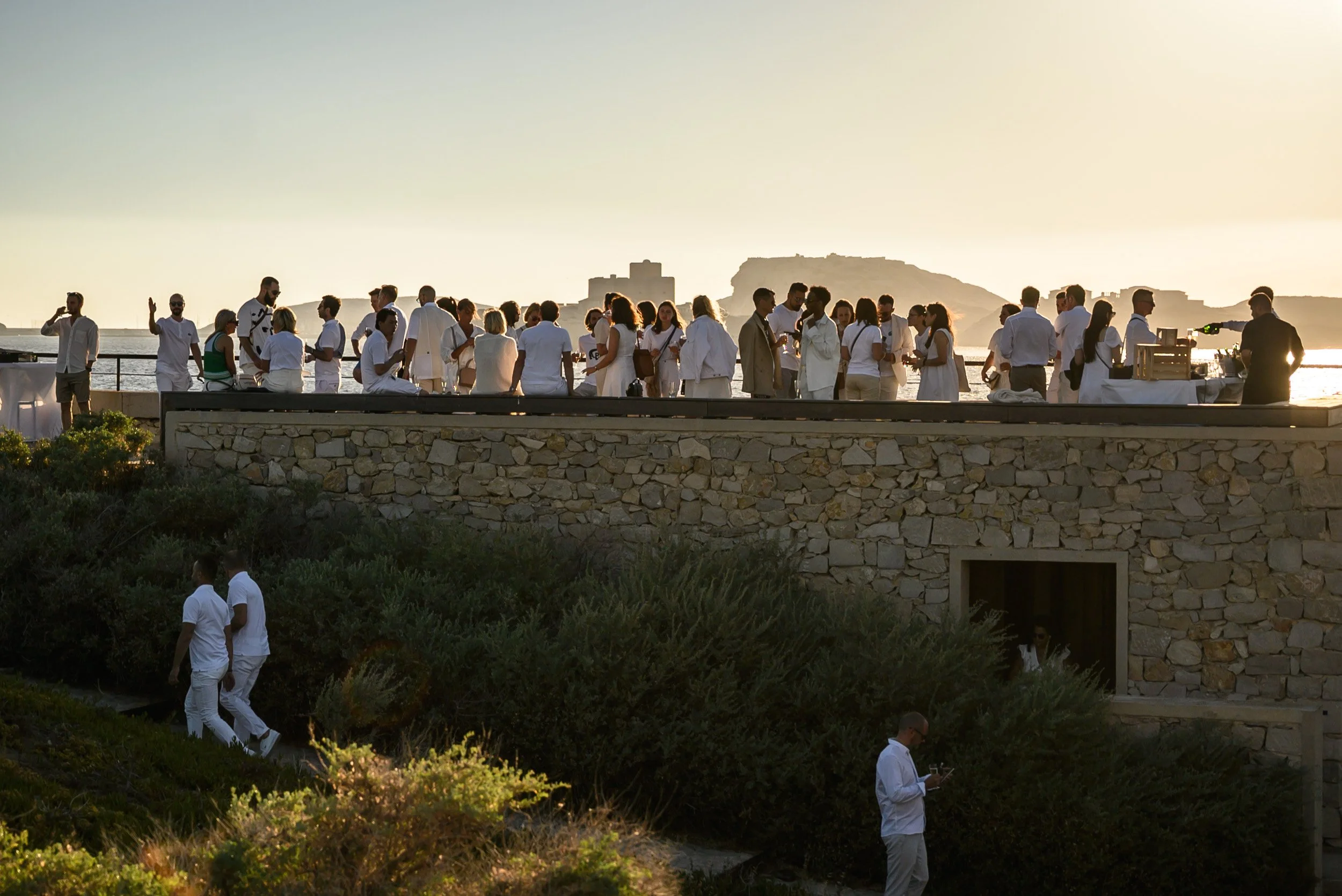 Groupe de personnes célébrant un événement en plein air au bord de la mer au coucher du soleil, avec un fort en arrière-plan.