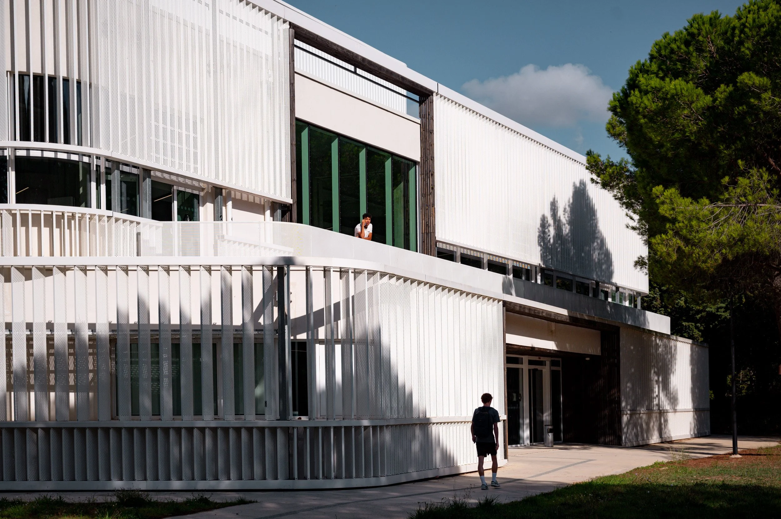 Un bâtiment moderne en blanc avec des grandes fenêtres en verre, entouré d'arbres verts, avec deux personnes dont une sur le balcon et une autre marchant en bas.