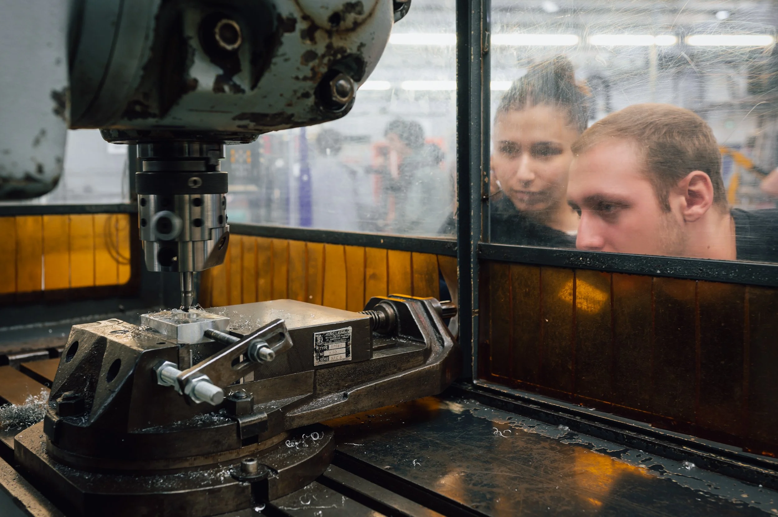 Deux jeunes hommes observer un outil de fabrication ou d'usinage à travers une vitre sale dans un atelier ou une usine.