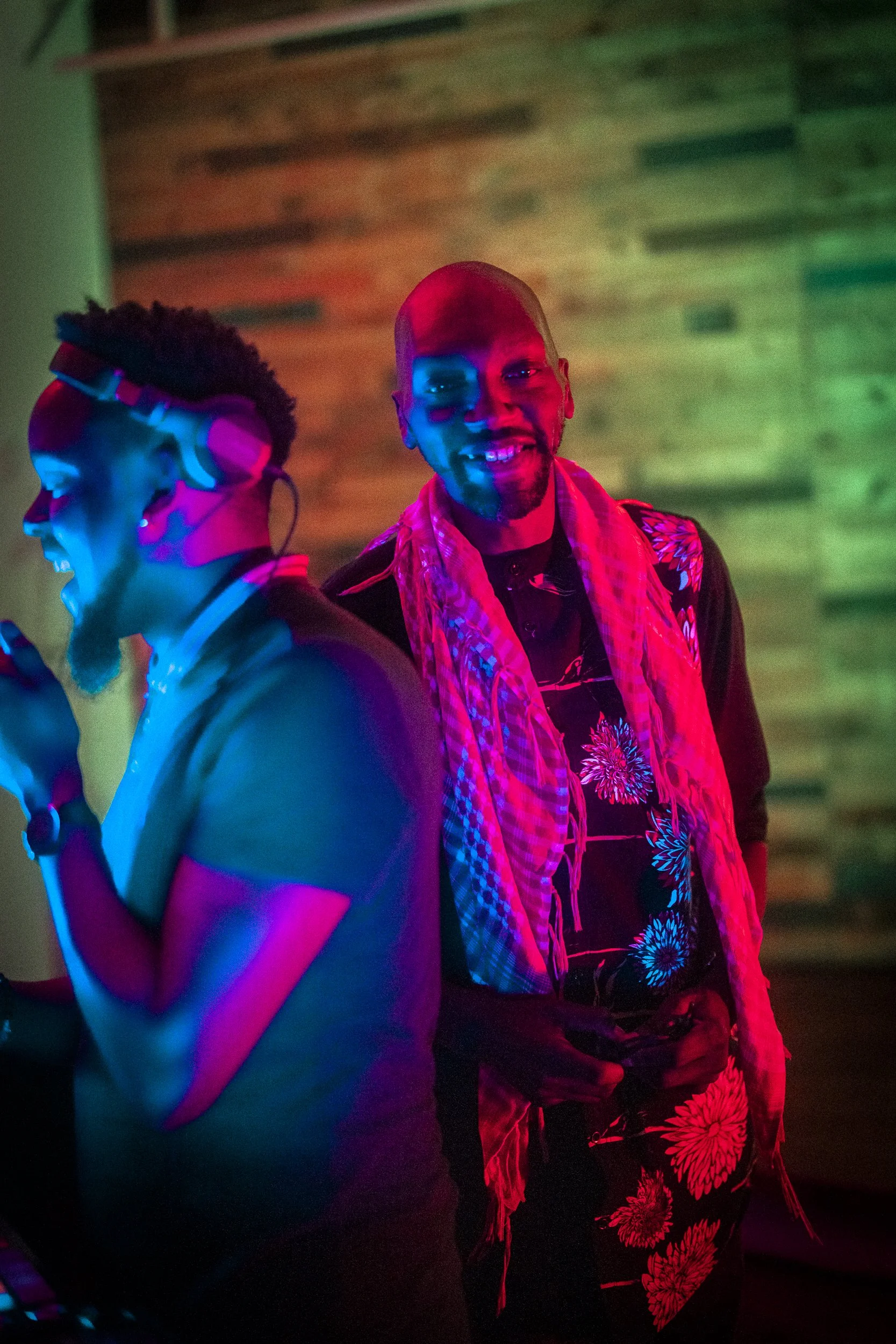Two men at a party, one with glasses and headphones smiling, the other bald with a patterned shirt and scarf, standing in front of a wooden wall, illuminated by colorful neon lights.