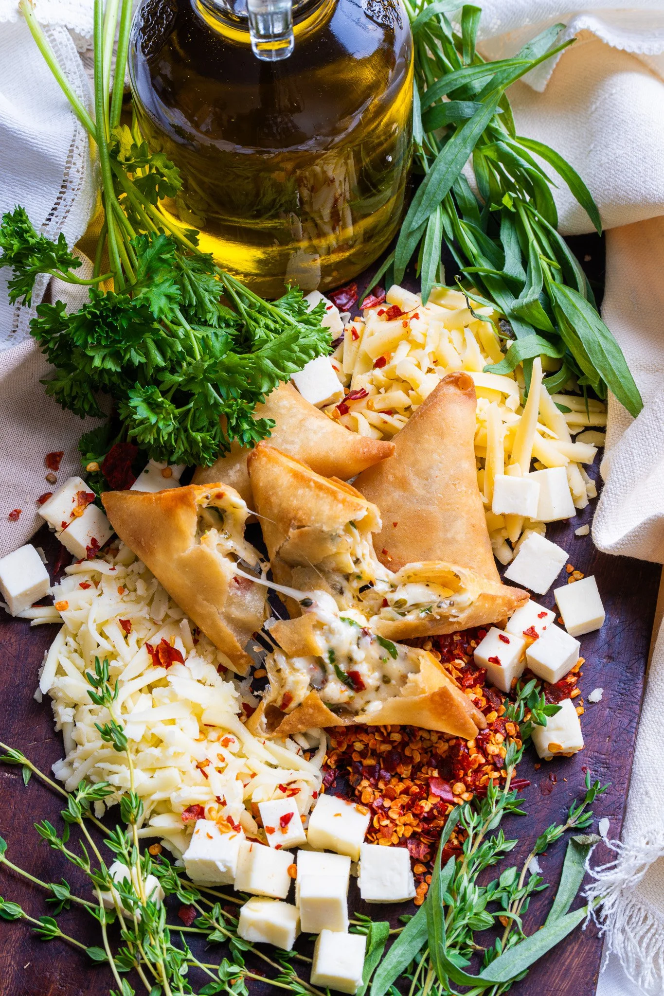 A variety of cheeses, fresh herbs, and spices around a small glass bottle of olive oil or vinegar.