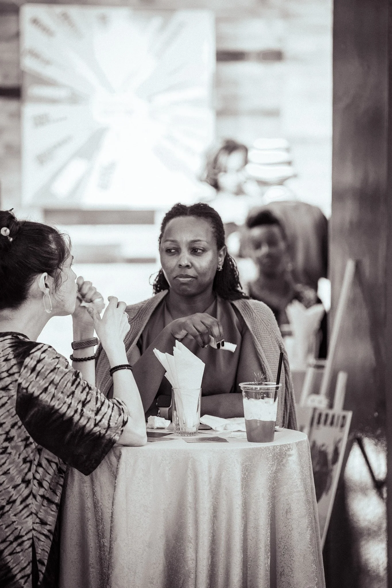 Two women are sitting at a table having a conversation in a coffee shop. One woman appears to be listening attentively while the other woman is speaking, with drinks and napkins on the table.
