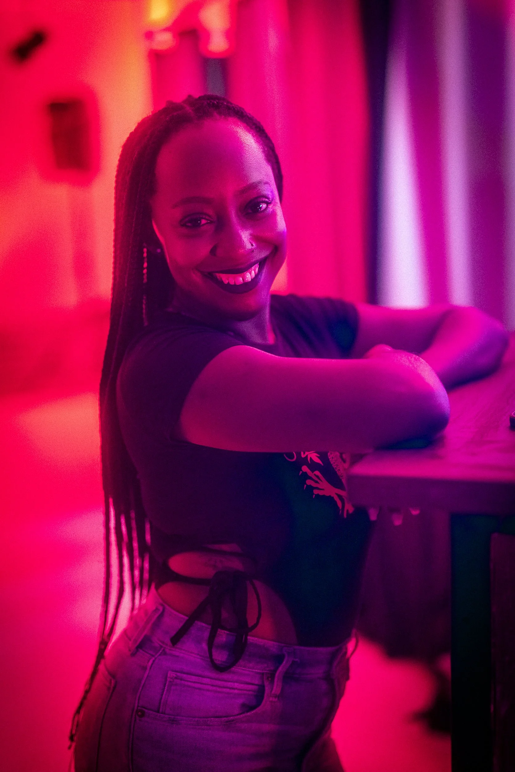 A woman with long dreadlocks smiling and leaning on a wooden surface, illuminated by pink and purple lighting.