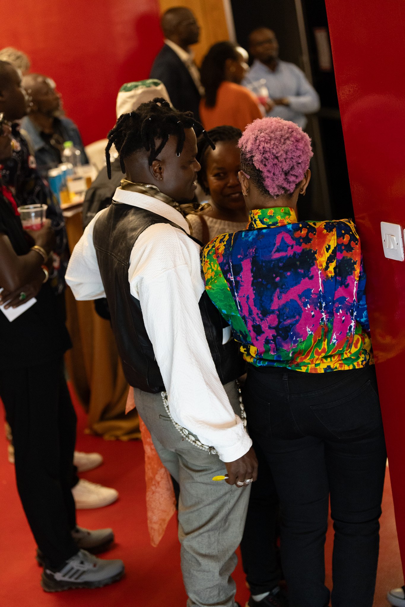 Two people with vibrant pink hair wearing colorful clothing are in a conversation near a red wall at a gathering of people.