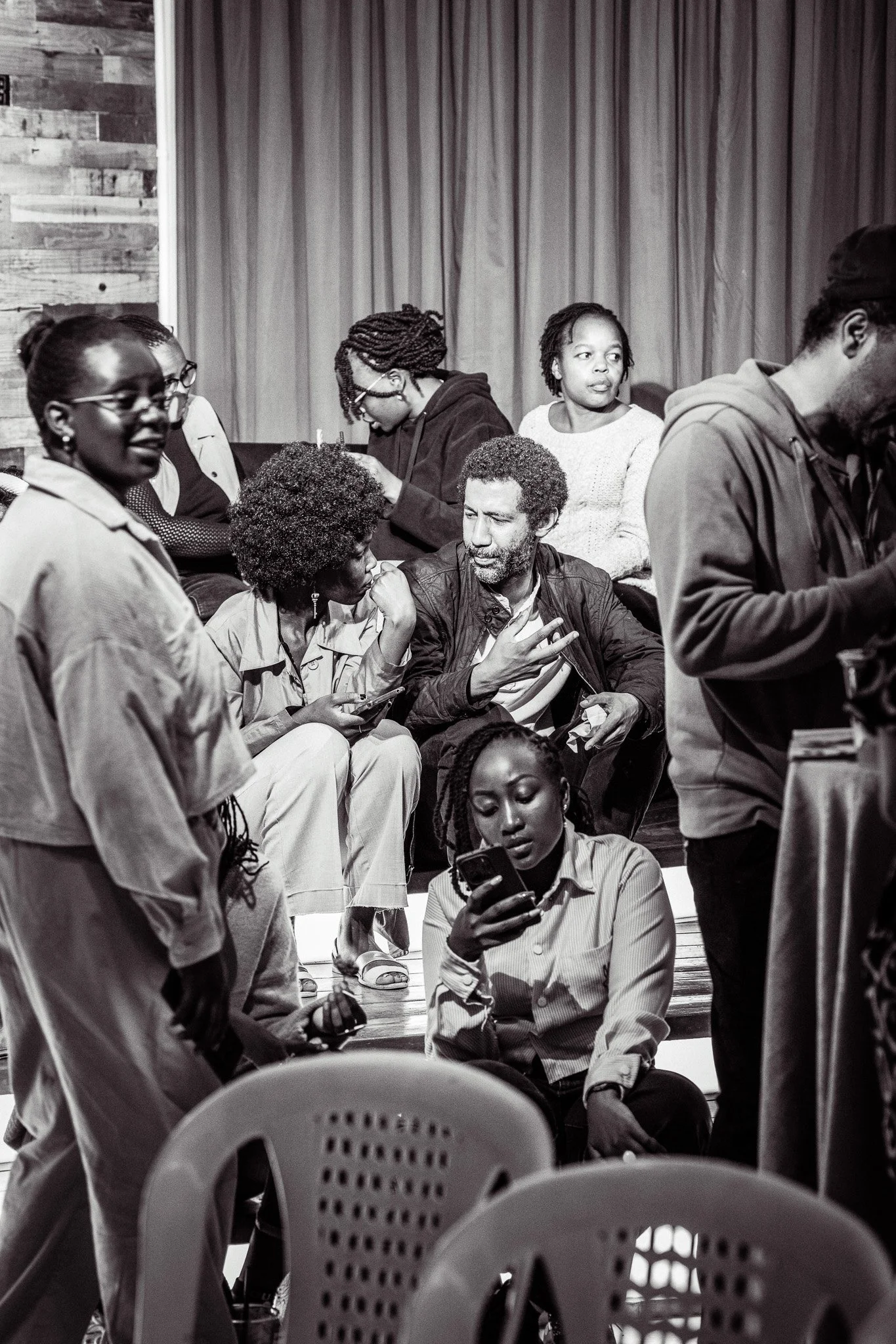 A group of people gathered indoors, some sitting and some standing, engaged in conversation and using their phones.