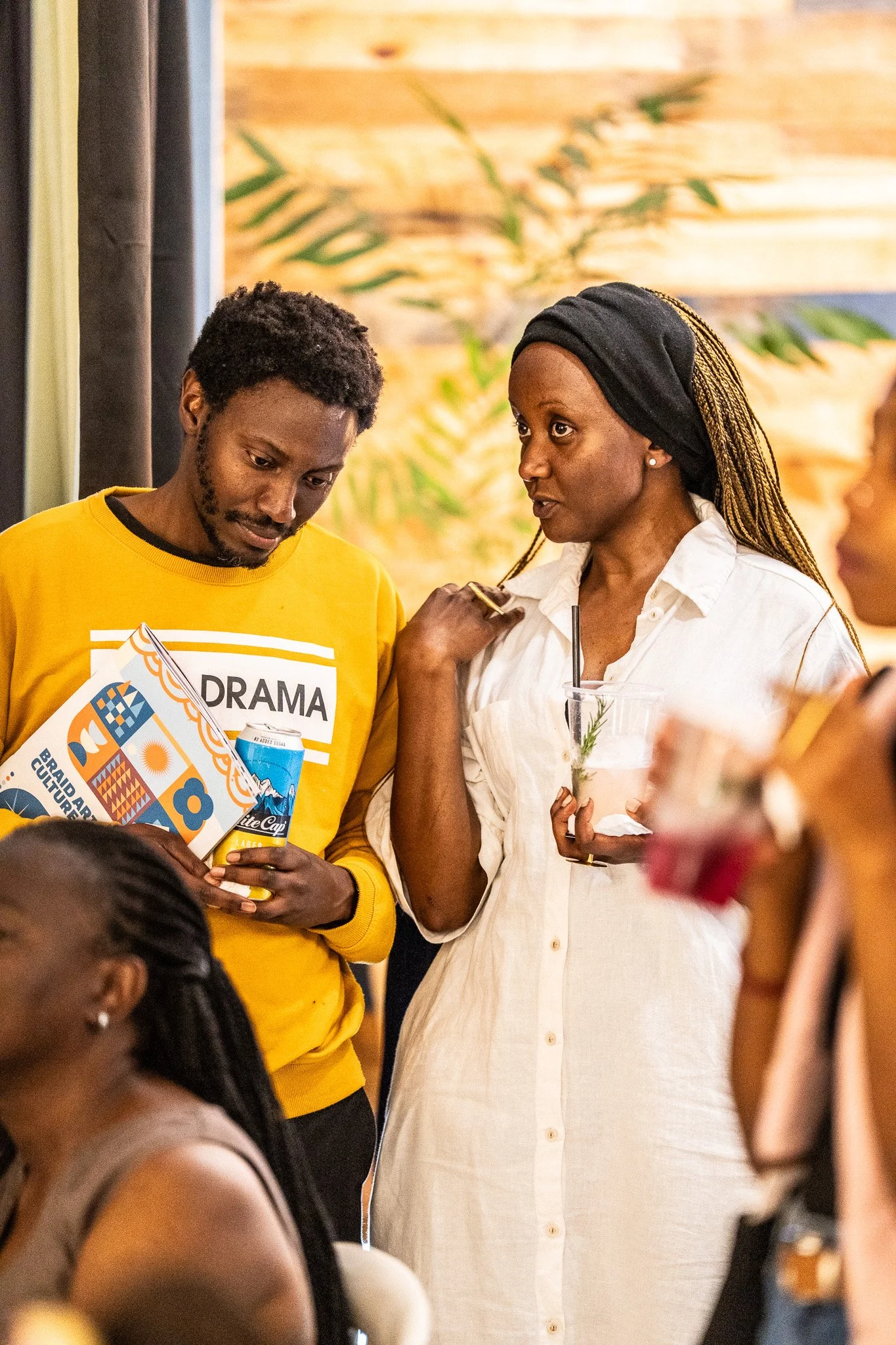 Two people in conversation at a social gathering, one wearing a yellow shirt and the other in a white dress, holding drinks.