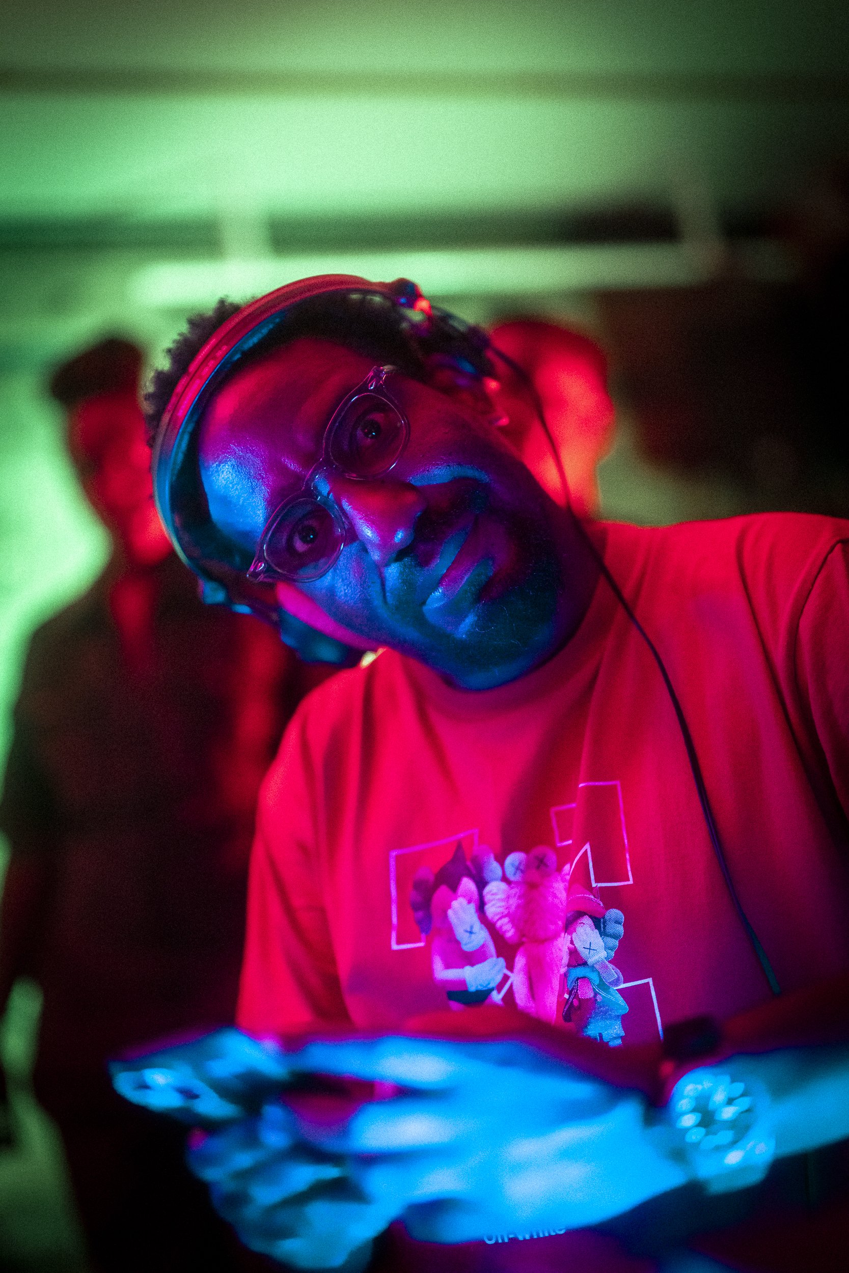 A man wearing glasses and a red t-shirt, illuminated by multicolored lights, looking at his phone, in a dimly lit environment with green and red lighting.