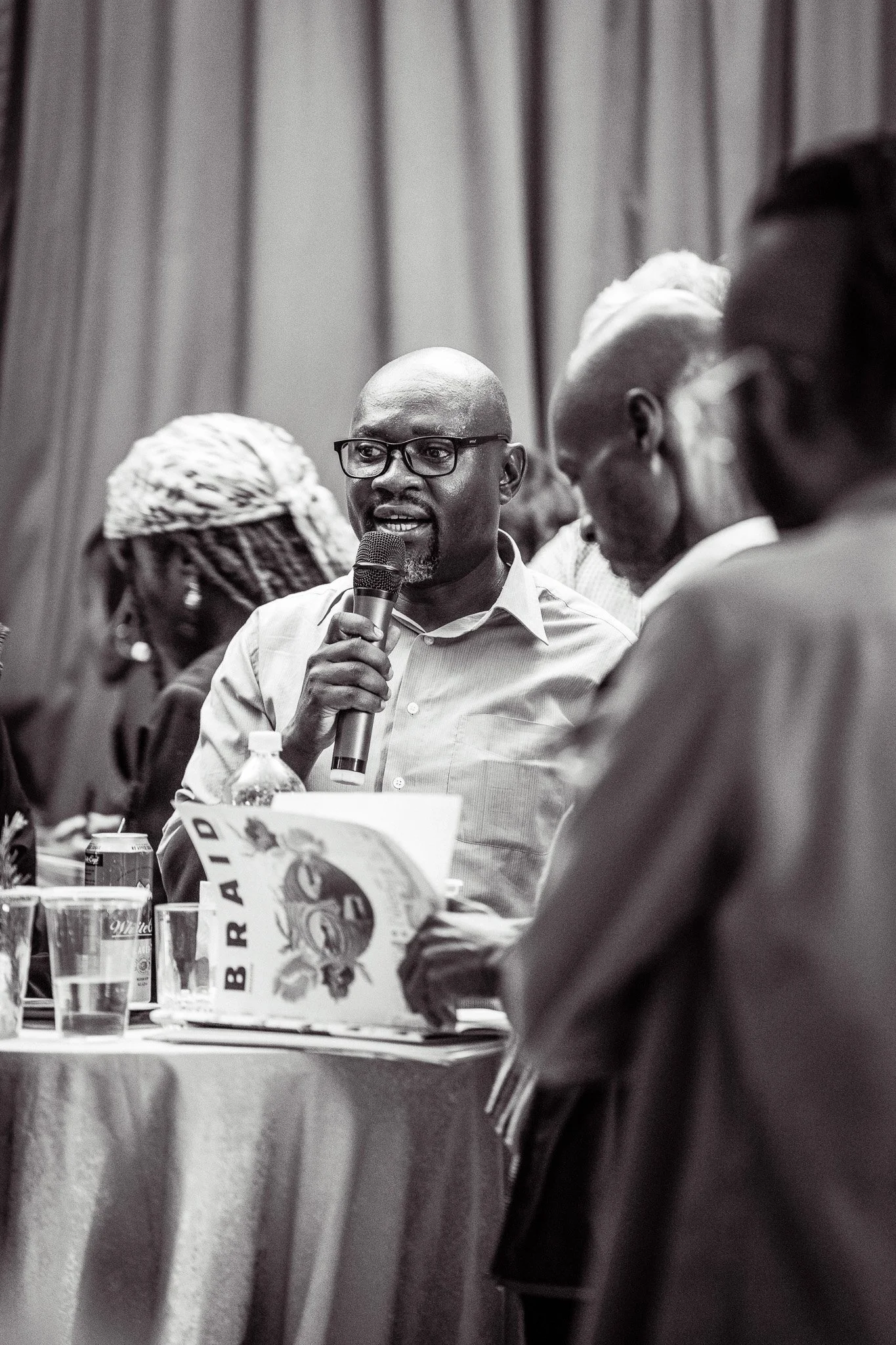 A man speaking into a microphone at a conference, with several people seated around a table in the background.