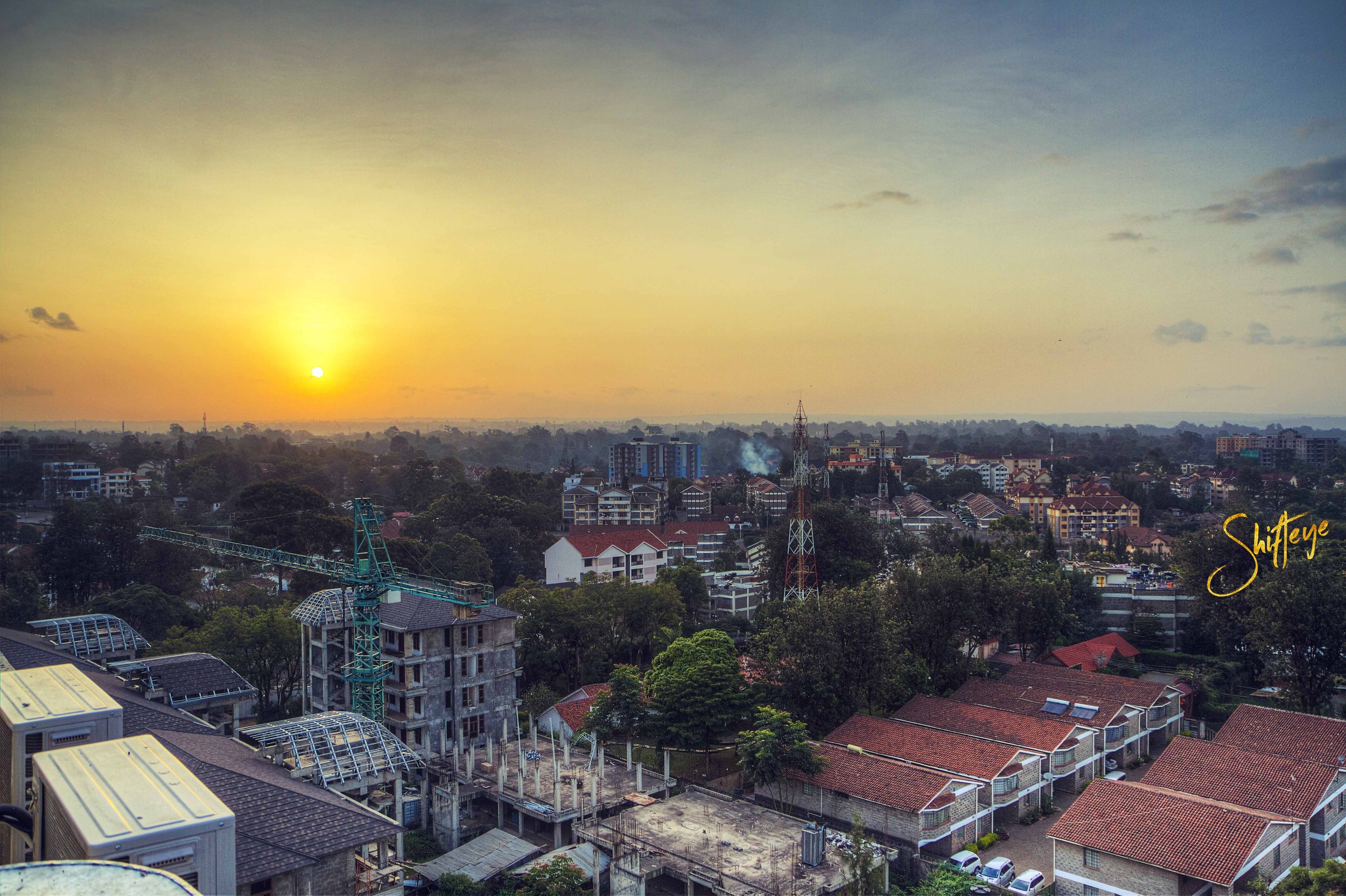 Aerial view of a cityscape featuring various buildings, a construction site with cranes, and a tower, under a setting sun creating an orange sky. Trees and residential rooftops are visible, with a partially clear sky.