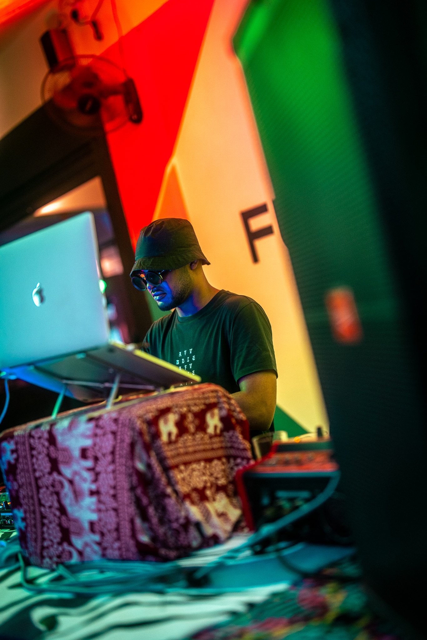 A DJ wearing sunglasses and a hat, working with equipment at a colorful, vibrant event, with rainbow background banners.