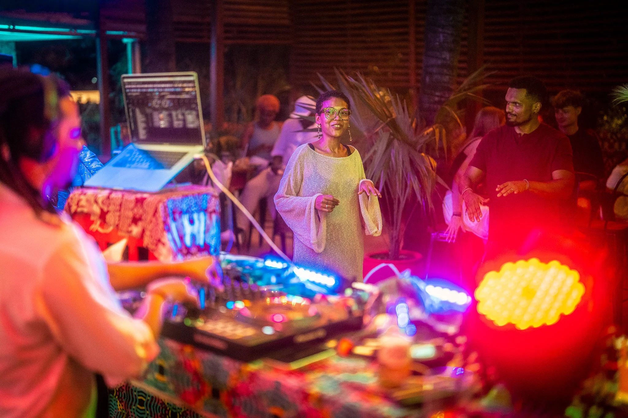 People dancing and socializing at a lively indoor party with colorful lights, a DJ playing music, and tropical plants in the background.