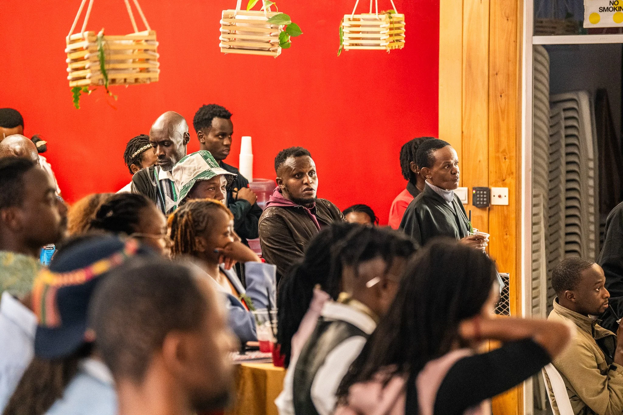 A group of diverse people attentively listening at an indoor event with a red wall and wooden accents.