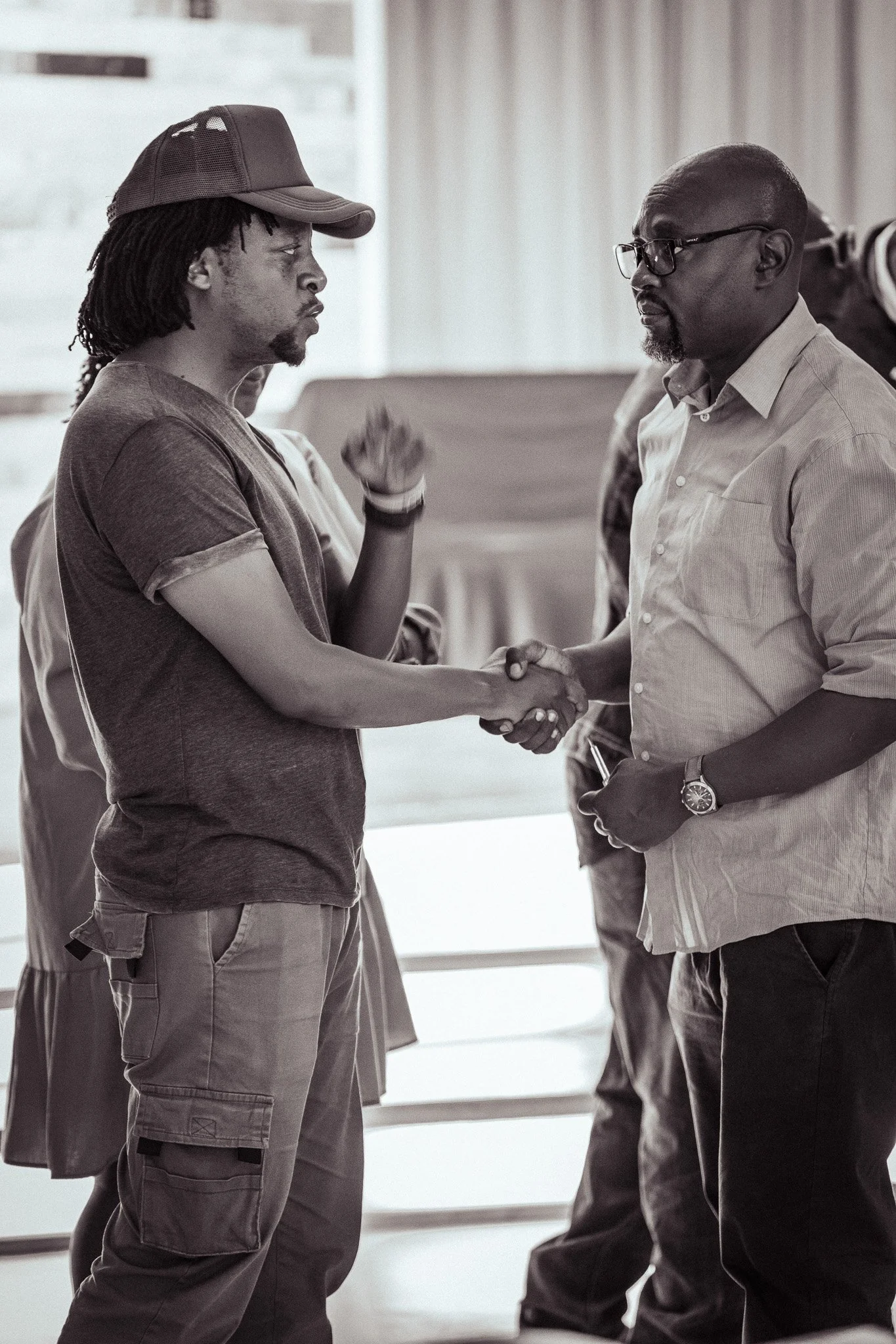 Two men shaking hands indoors, one wearing a baseball cap and the other wearing glasses, with a blurred background of other people.