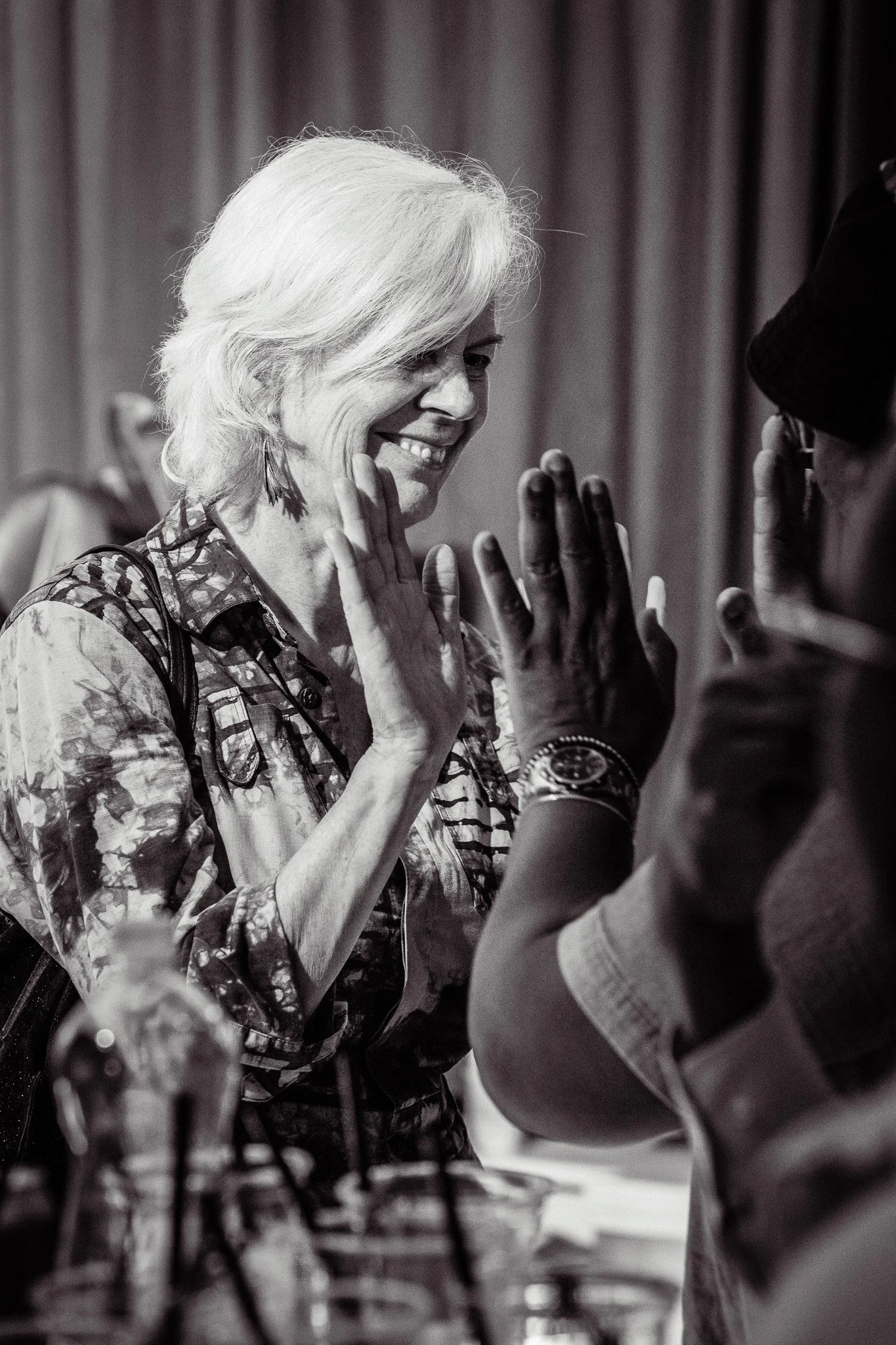 A black and white photo of a woman with short, wavy white hair smiling and raising her hand to high-five a man wearing glasses and a hat. There are drinks on the table in front of them.