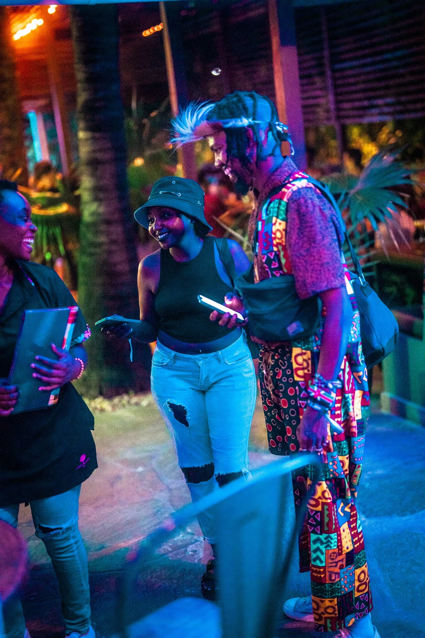Three people having a conversation at a lively, colorful party or event, illuminated by neon lights and surrounded by tropical plants.