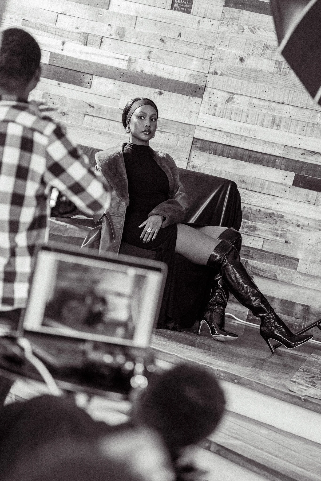 A woman sitting on a black couch in a photoshoot, wearing a black dress and thigh-high leather boots, with a fur-lined jacket. She poses confidently against a wooden wall background while photographers and a monitor record the scene.
