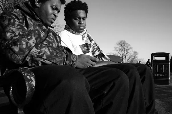 Two young boys sitting on a bench outdoors, looking at a phone.