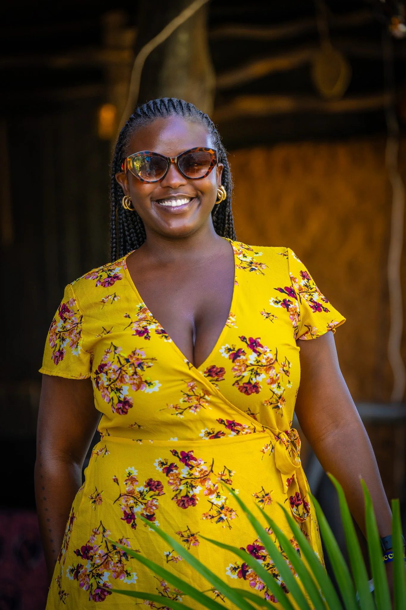 A woman wearing a yellow floral dress and sunglasses, smiling outdoors with greenery in the foreground.