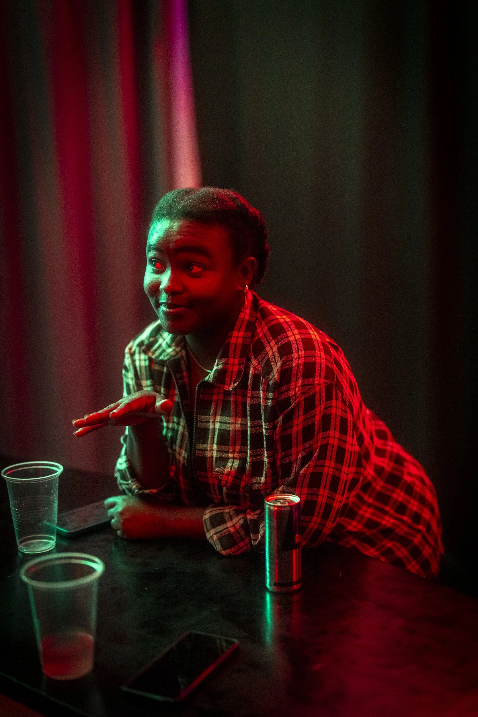 A woman with dark skin and short braided hair is sitting at a table in a dimly lit room with colorful lighting, wearing a red and black plaid shirt. She is leaning on the table with one arm, while the other hand is raised with her palm down, as if in