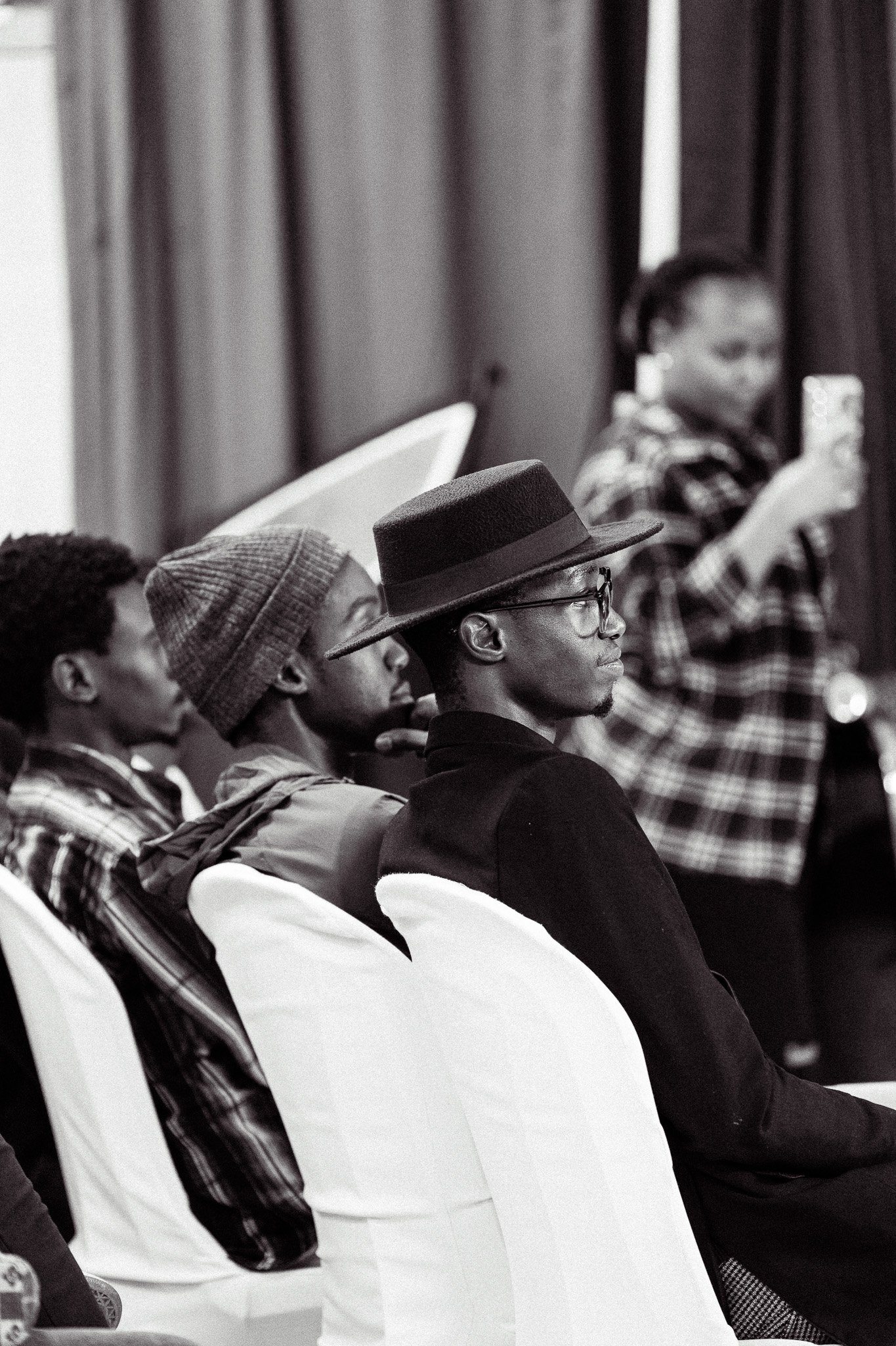 Black and white photo of four people sitting in chairs, listening attentively at an event. A woman in the background, standing, is taking a photo with her phone.