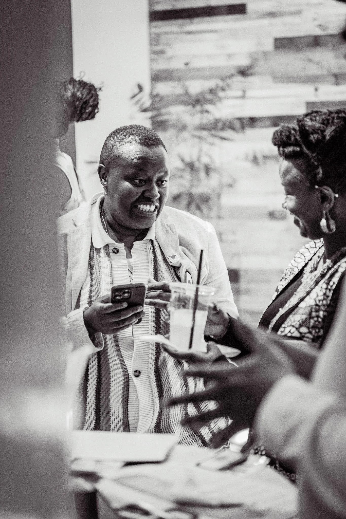 Group of women socializing indoors, with one woman showing something on her phone to others, in a setting that appears to be a cafe or restaurant.