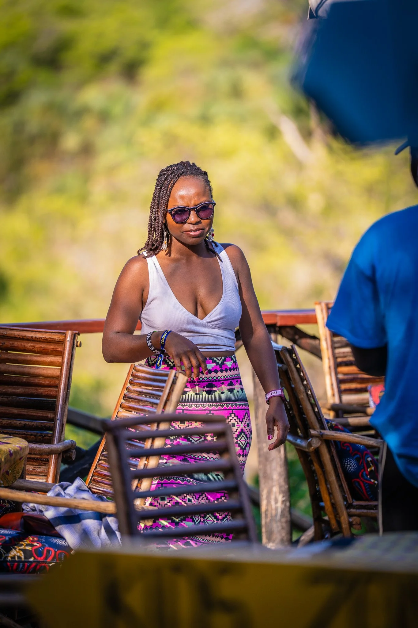 A woman wearing sunglasses and a white tank top stands outdoors among wooden chairs, with a blurred green background, engaged in conversation with a man.
