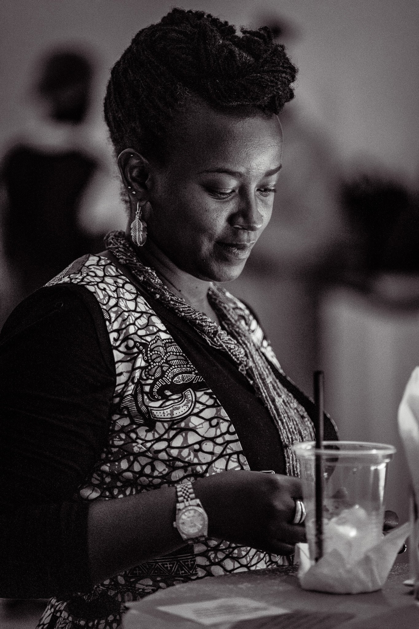 Black and white photo of a woman with short, styled hair, wearing earrings and a patterned shirt, sitting at a table with a glass of iced drink with a straw and some food, looking down.