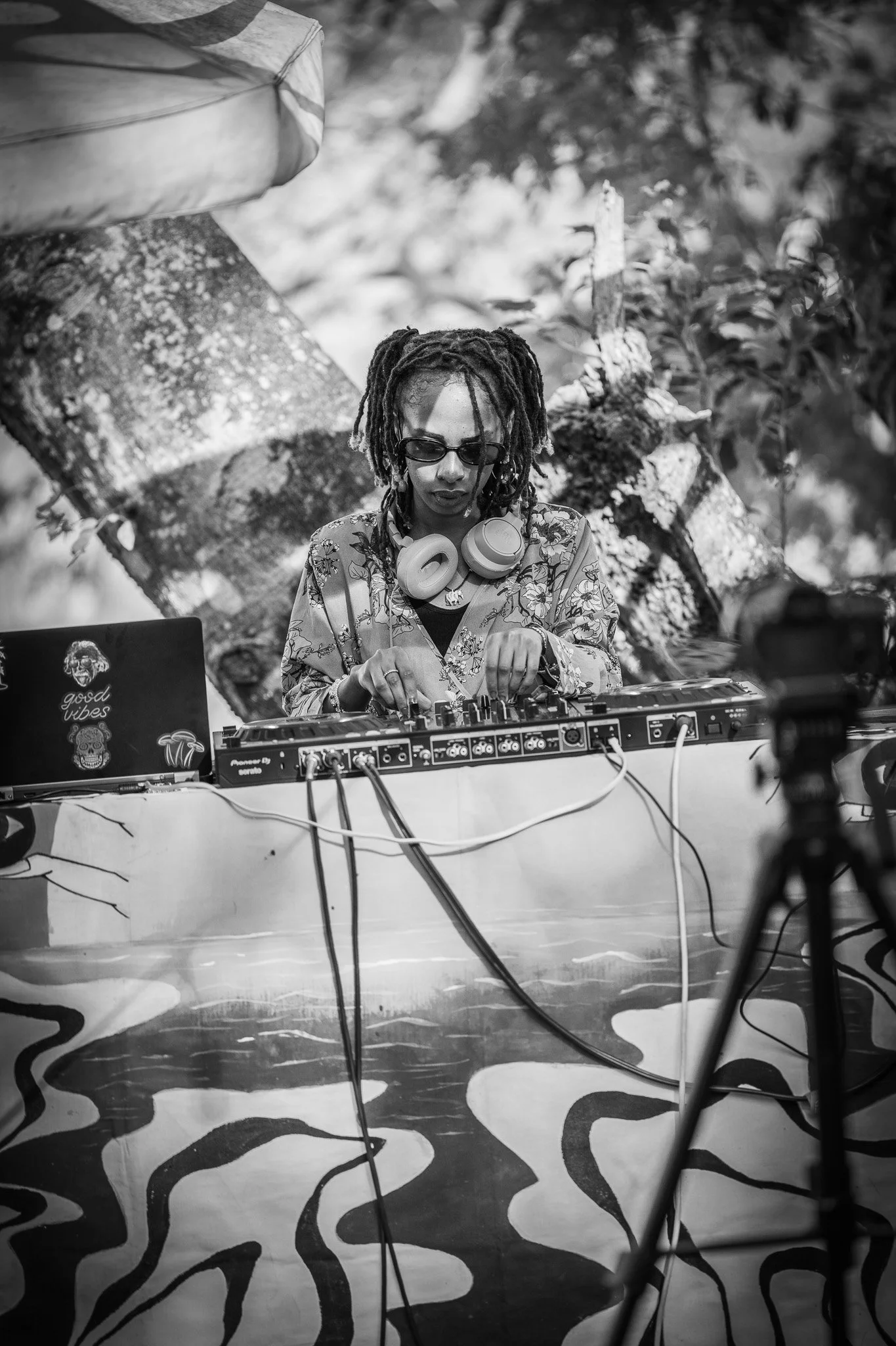 A female DJ with dreadlocks, sunglasses, and headphones around her neck, performing outdoors behind a DJ setup, in black and white.
