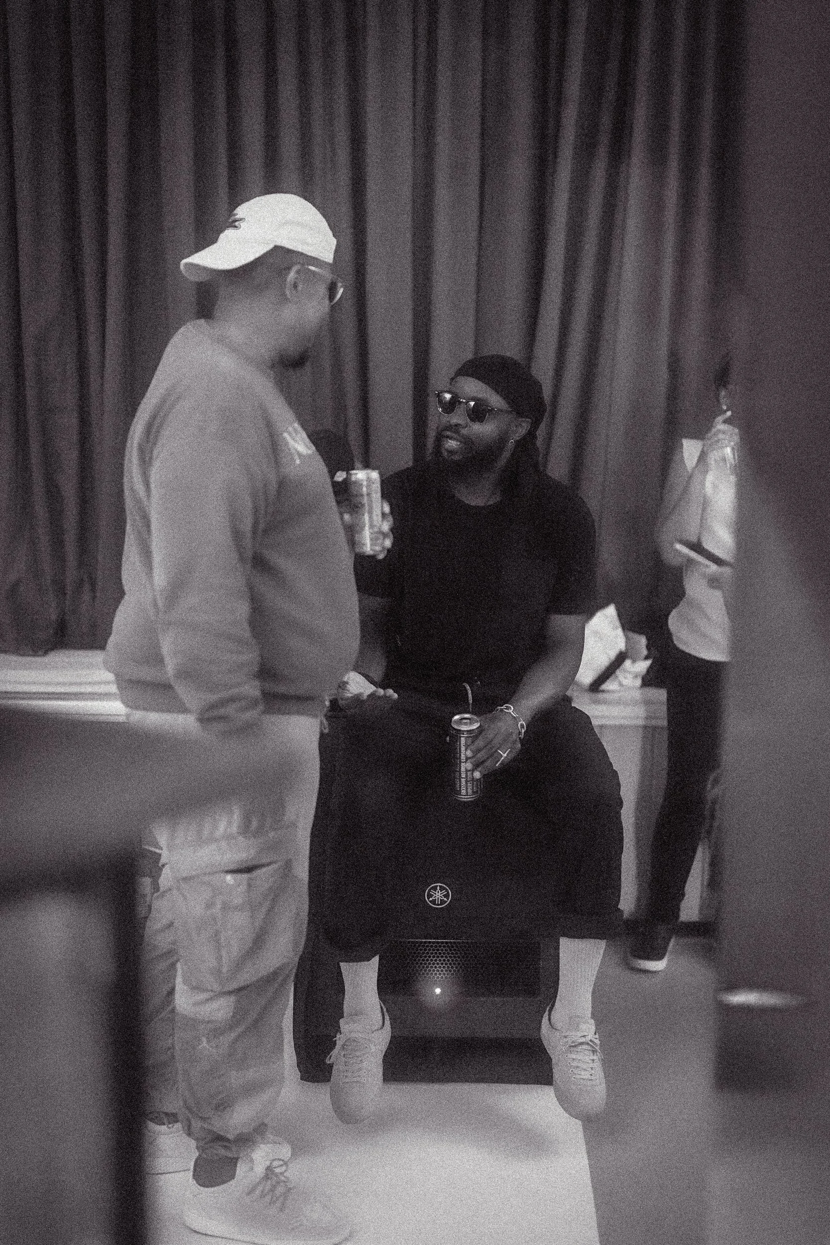 Two men engaged in conversation, one standing with his back to the camera wearing a baseball cap and sunglasses, the other sitting on a speaker wearing sunglasses and a beanie, both holding canned drinks, in front of dark curtains.