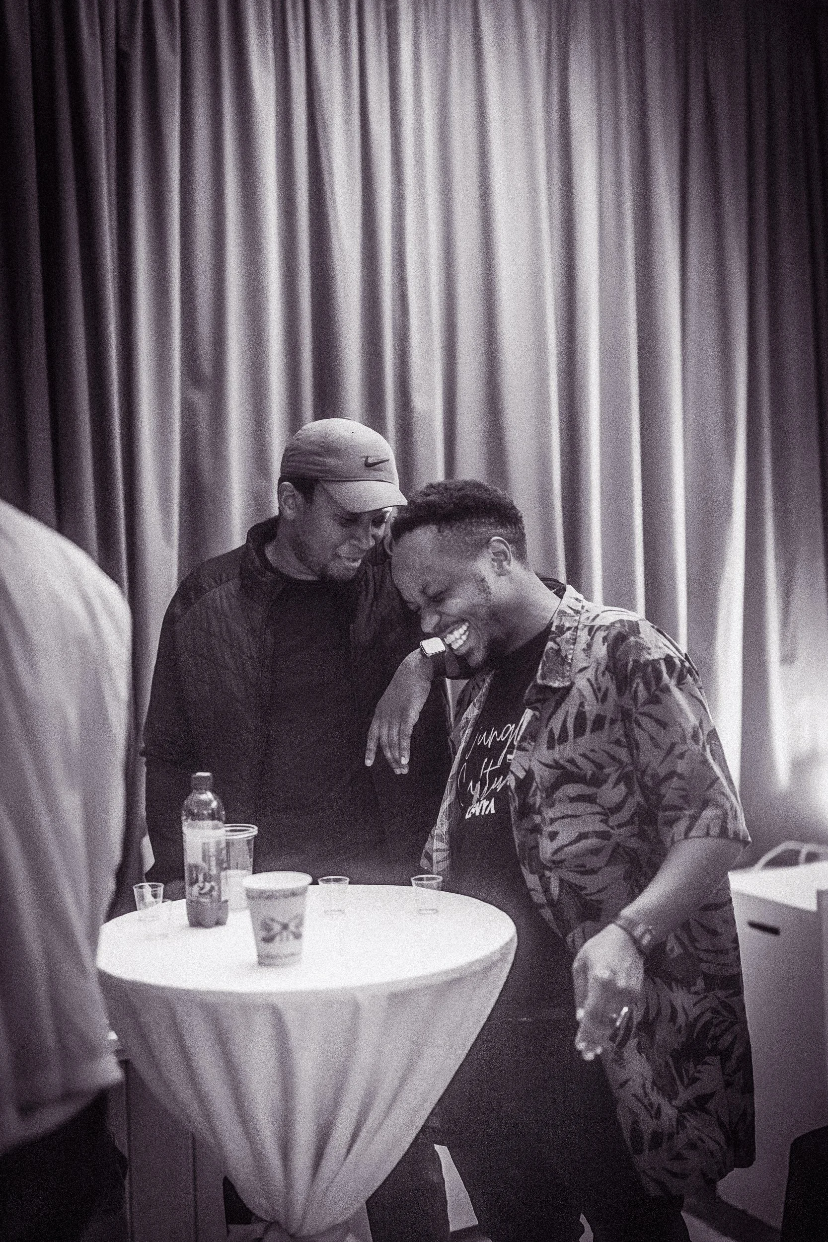 Two men smiling and leaning their heads together at a round table with drinks, with curtains in the background.