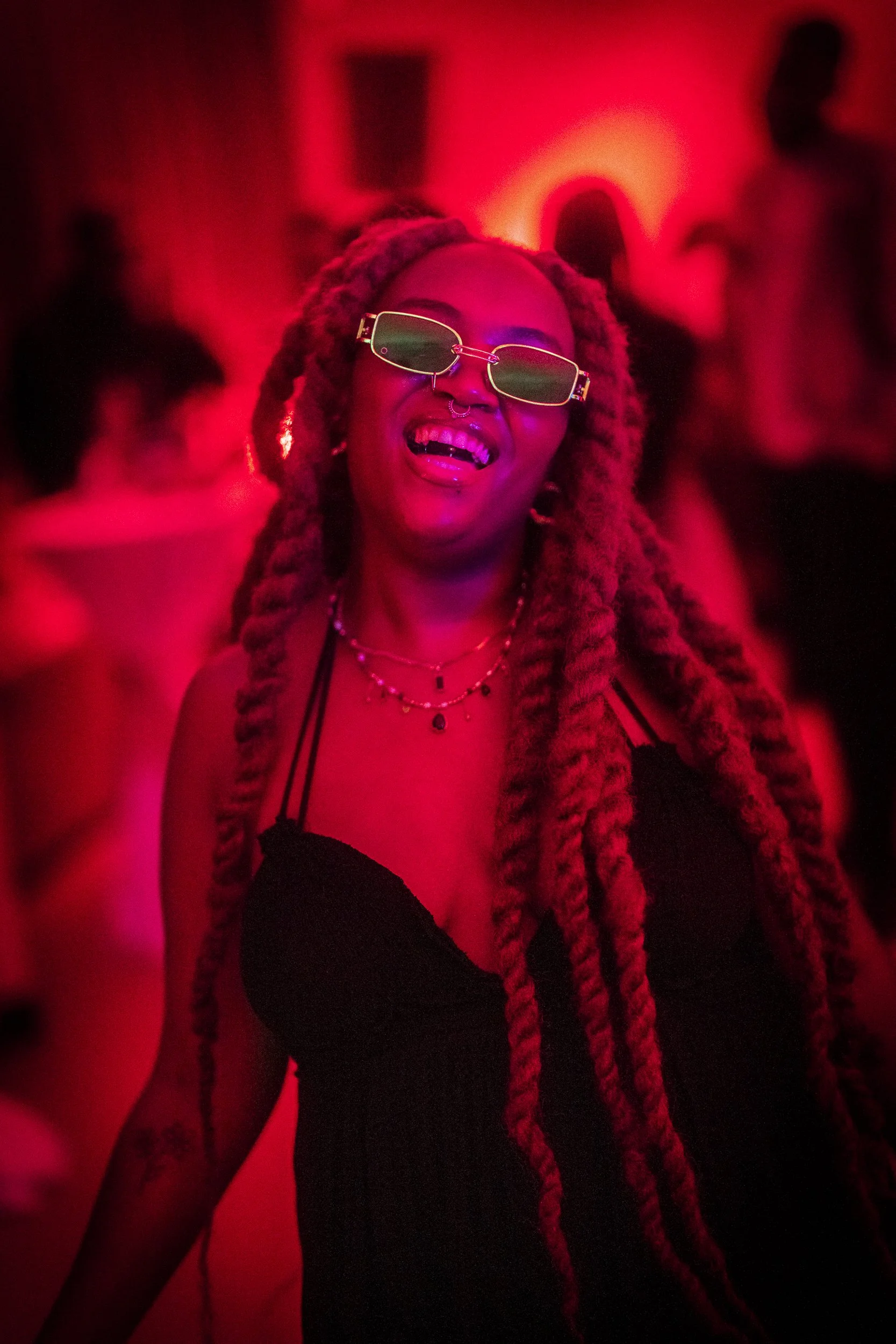 A woman with long, braided hair dancing at a party, wearing sunglasses, a black dress, and layered necklaces, illuminated by red lighting.