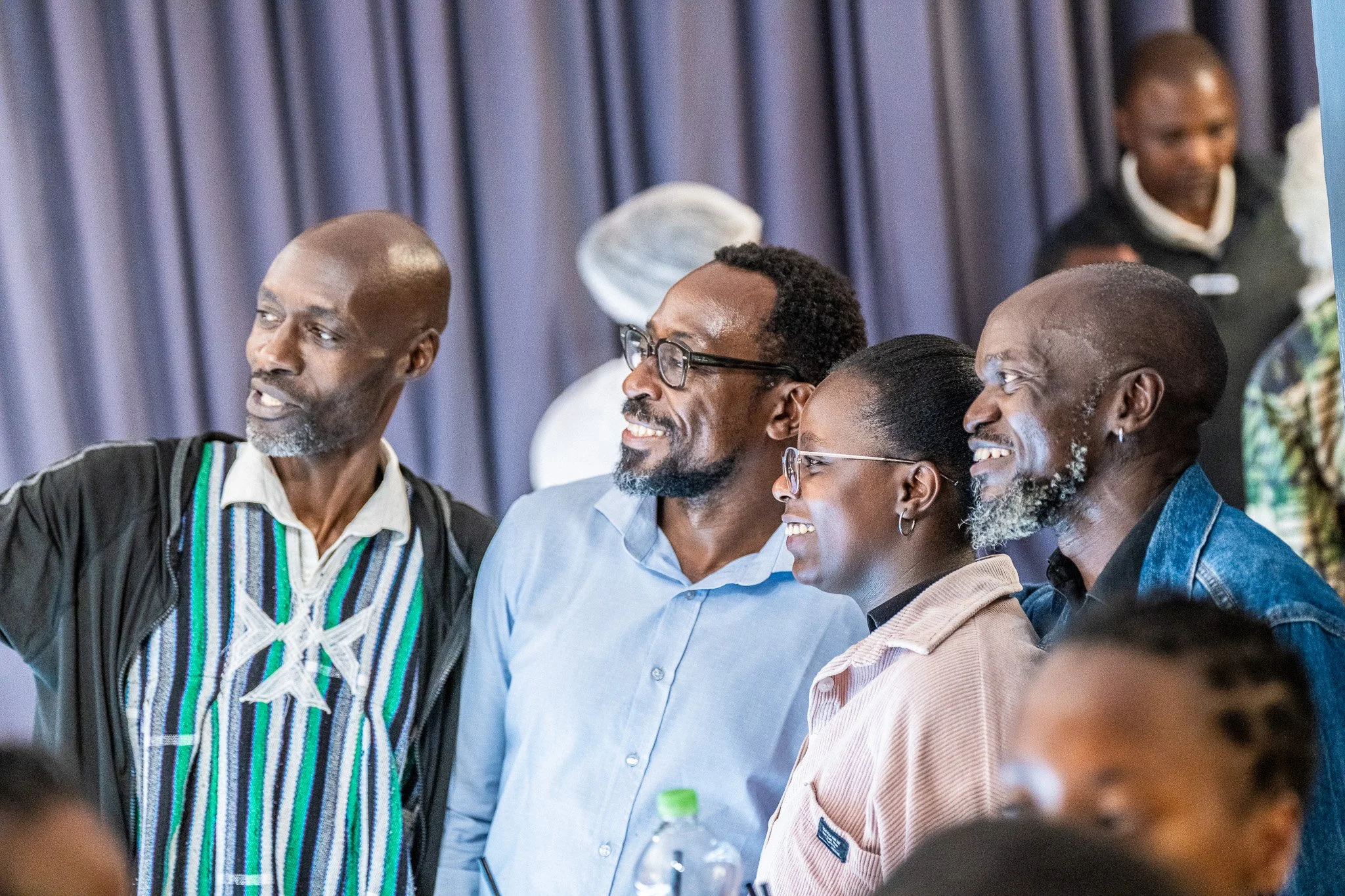 Four people standing in a row, smiling and engaging with each other, in a setting with blue curtains in the background.