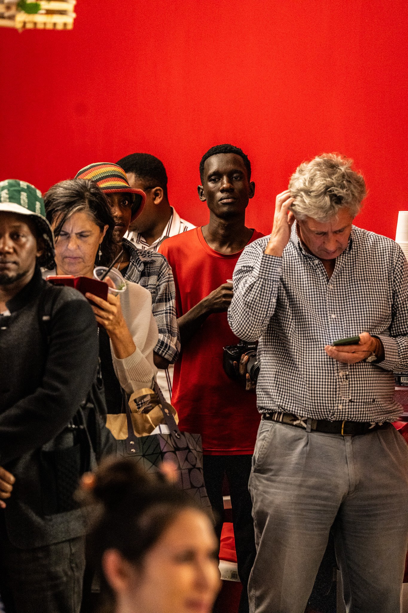 A diverse group of people standing in a line against a red background, looking at their phones or waiting.