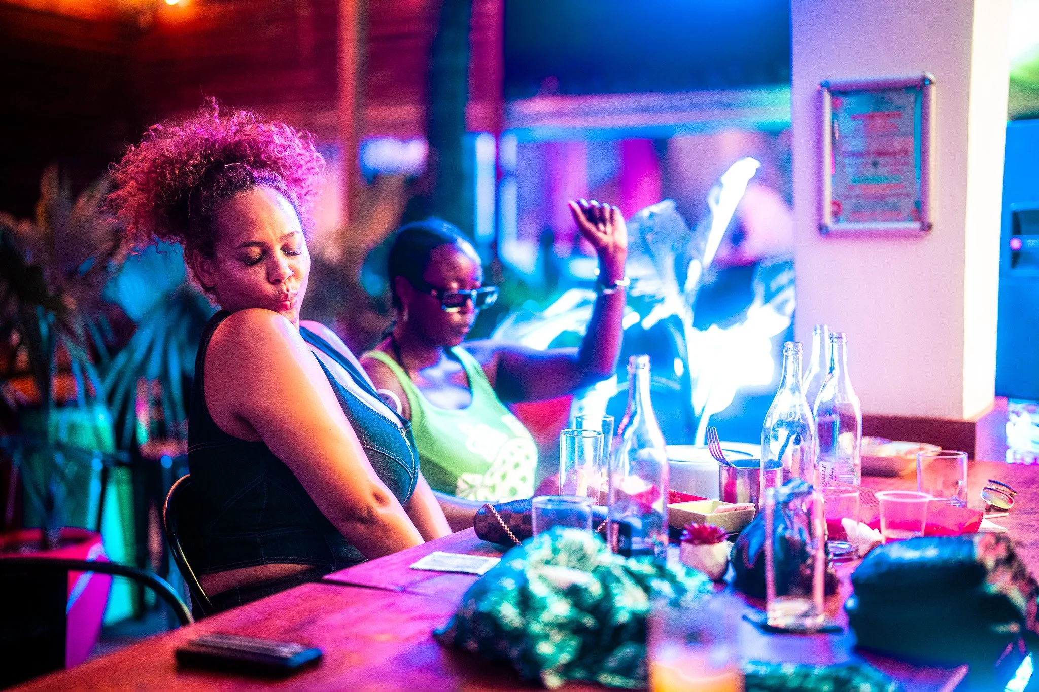 Two women dancing and enjoying themselves at a bar or club with colorful lights and empty tables with bottles and glasses.
