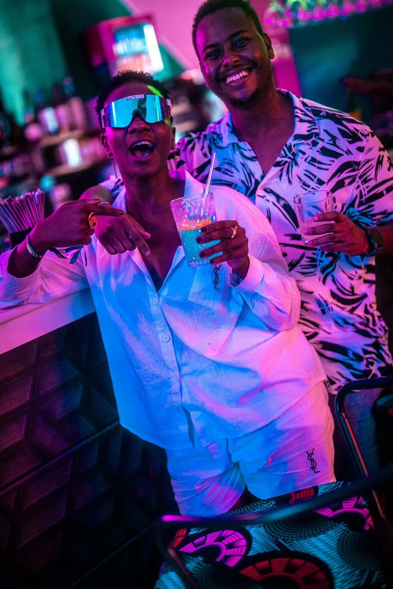 Two people enjoying a night out at a bar, smiling and holding drinks, with vibrant neon lighting in the background.