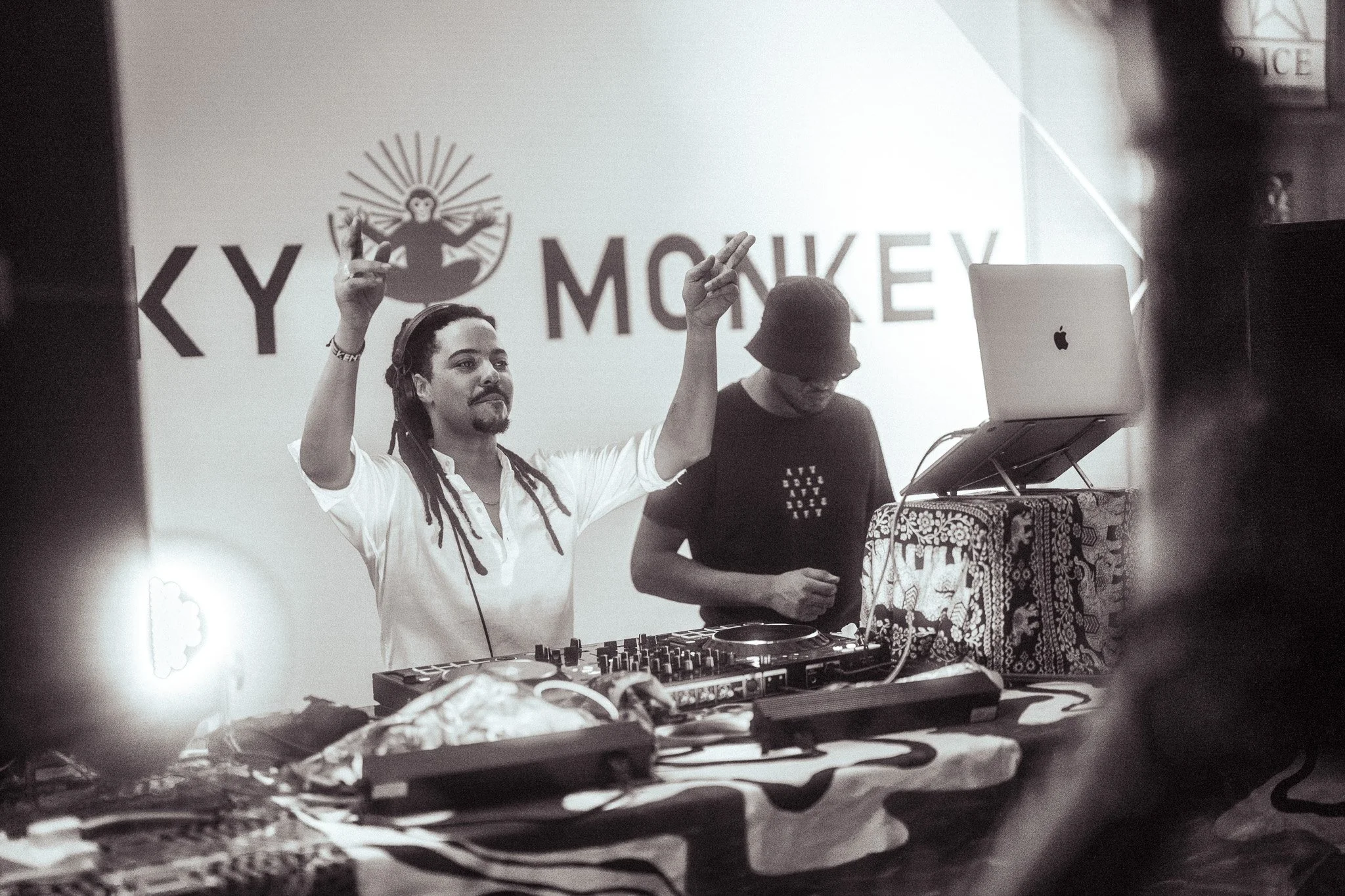 A DJ with dreadlocks, wearing headphones and a white shirt, is raising one hand with two fingers up at a DJ booth. Beside him, another person is looking down at equipment. There is a large sign with a monkey and the words 'KEY MONKEY' behind them.