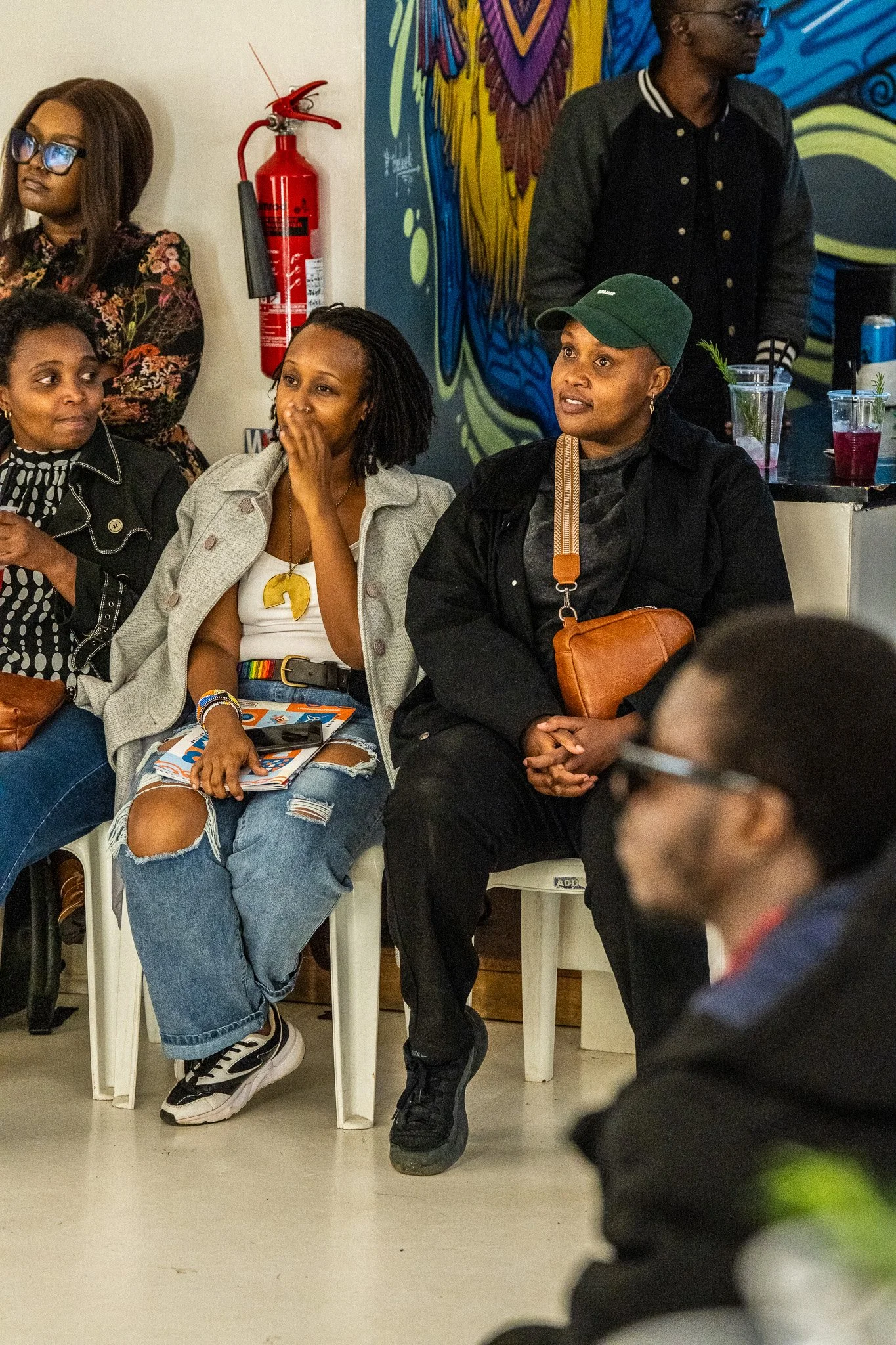 Group of black women sitting and listening at an indoor event, with colorful mural art in the background.