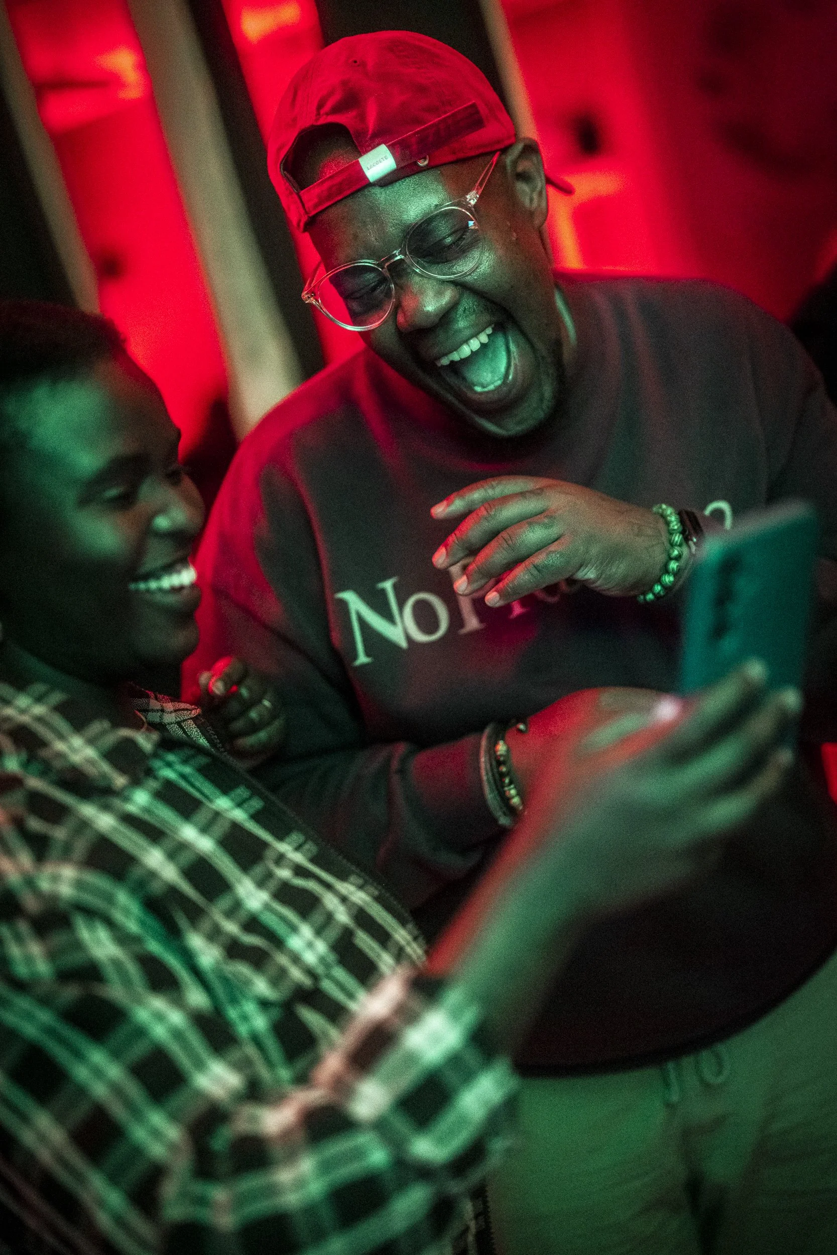 Two people smiling and laughing while looking at a smartphone in a dimly lit room with red and green lighting.