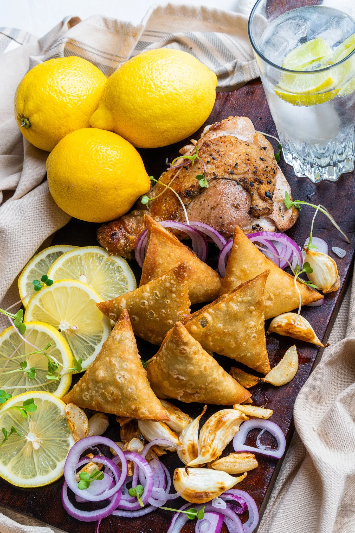 Lemon halves, whole lemons, grilled chicken, fried samosas, sliced red onions, and garlic cloves on a wooden board with a beige cloth in the background.